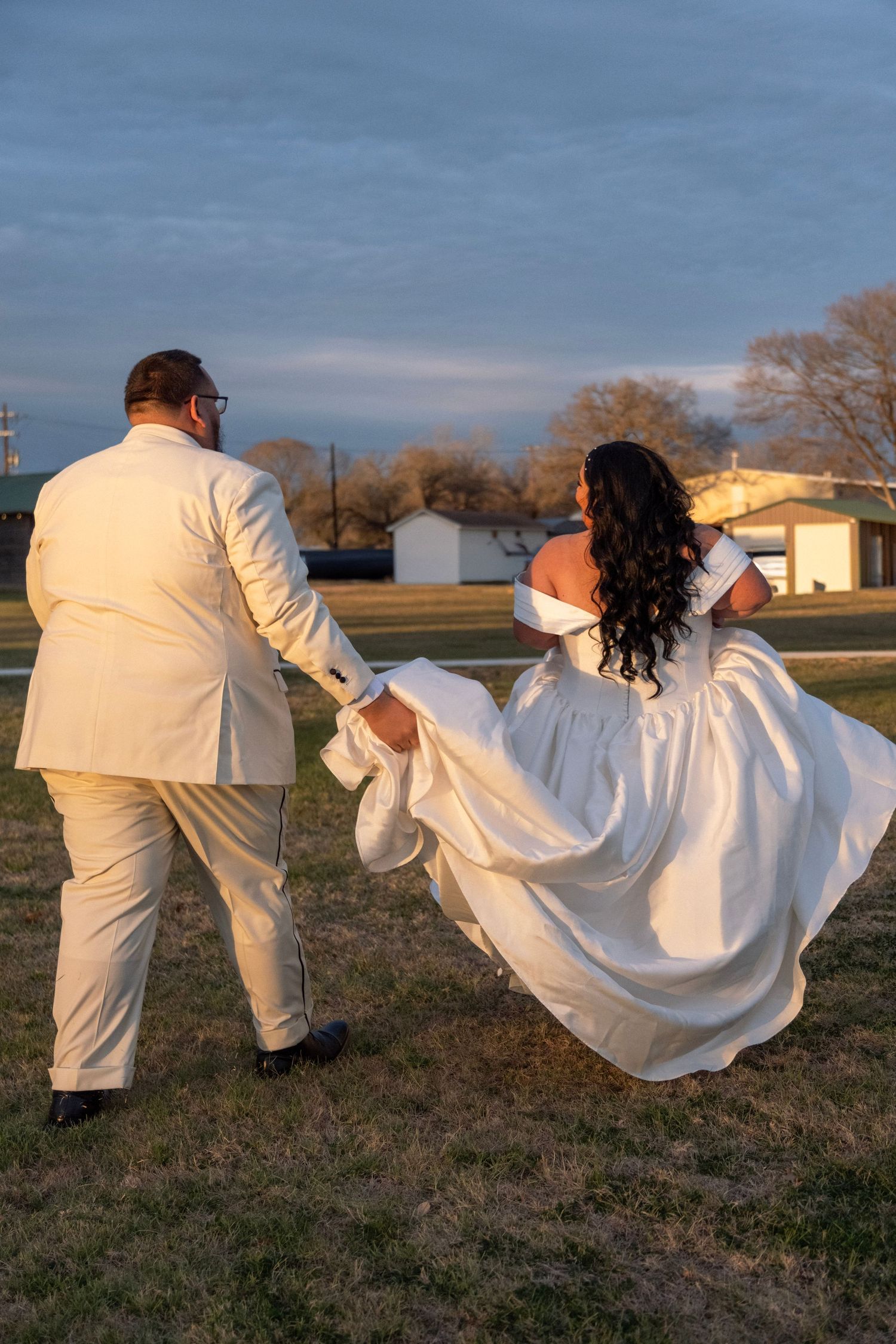 Bride and groom walk hand in hand across field at golden hour, wedding dress flowing in wind.
