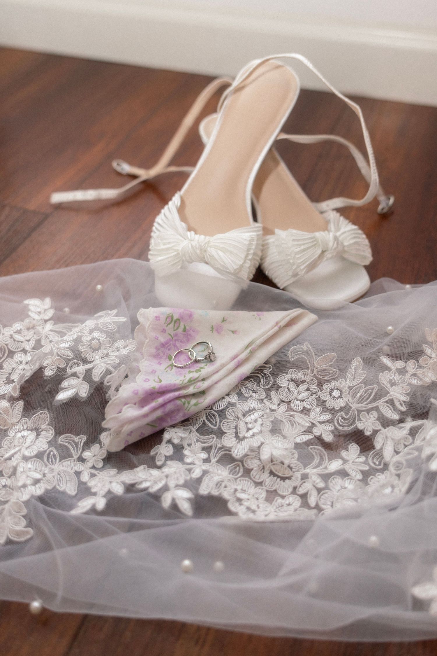 White bridal heels with bow detail resting on lace veil with floral handkerchief and wedding rings.