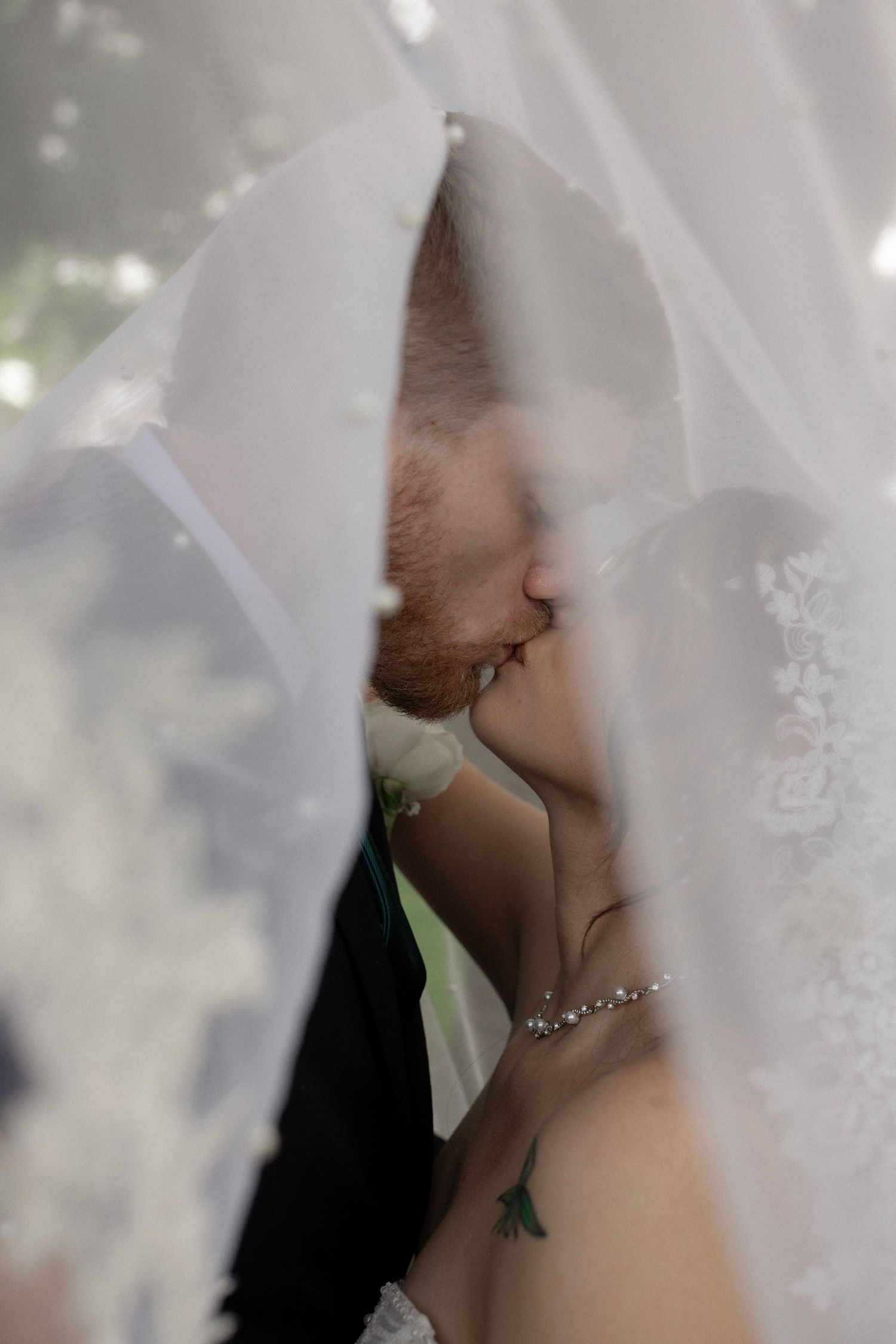 Bride and groom share a kiss hidden beneath a flowing white lace wedding veil outdoors.