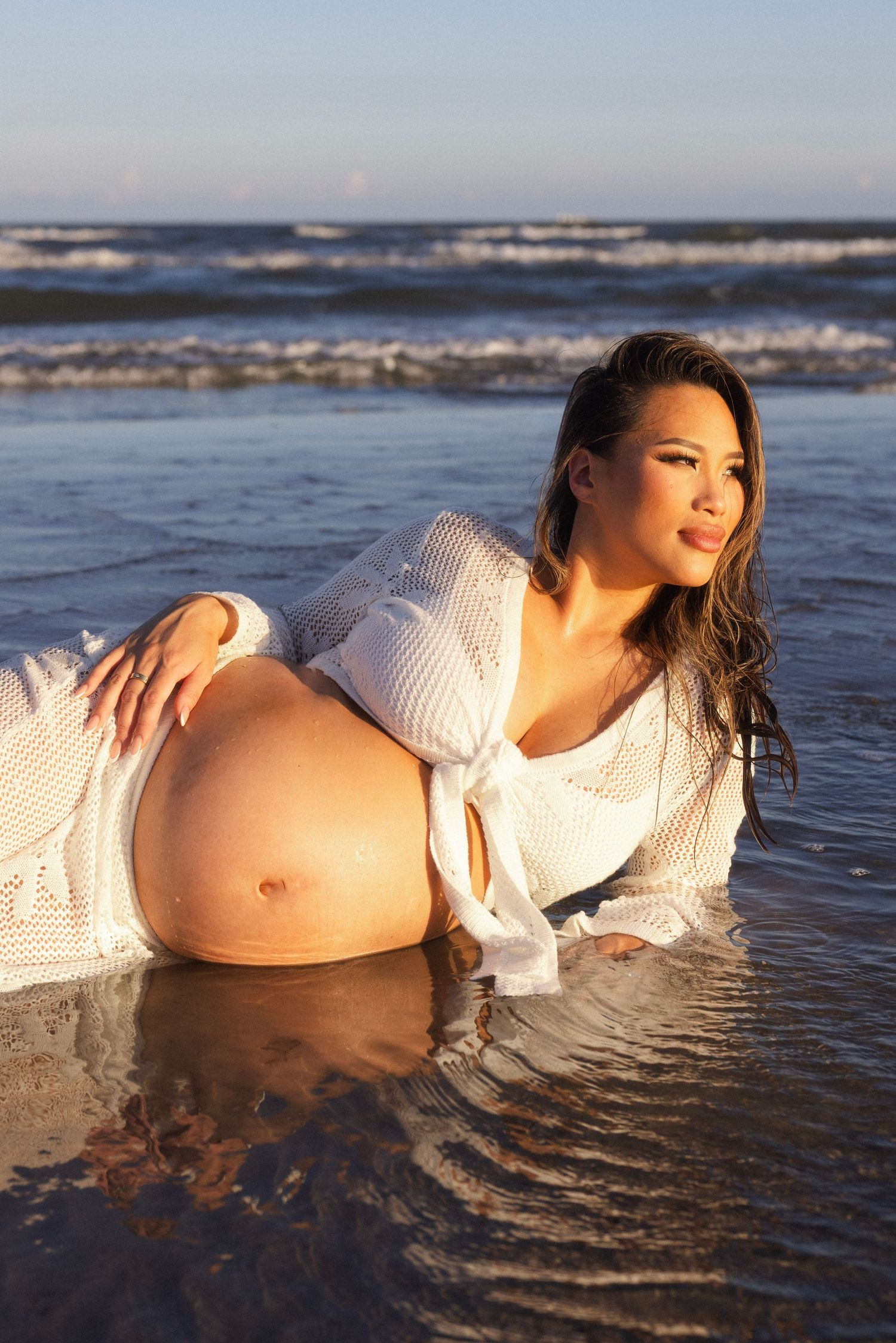 Pregnant woman in white knit dress posing in shallow ocean water during golden hour at the beach.