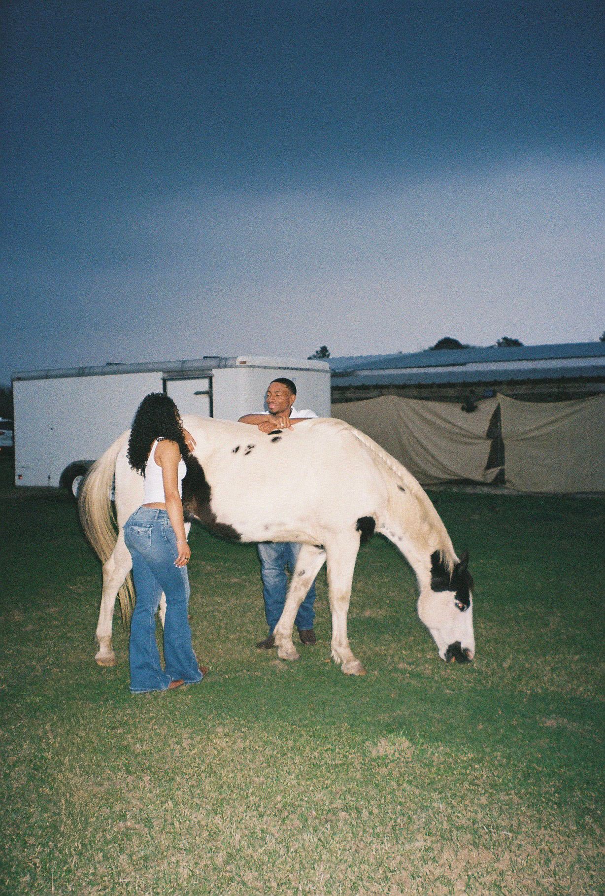 Couple laughing together beside a grazing black and white horse at dusk outdoors.