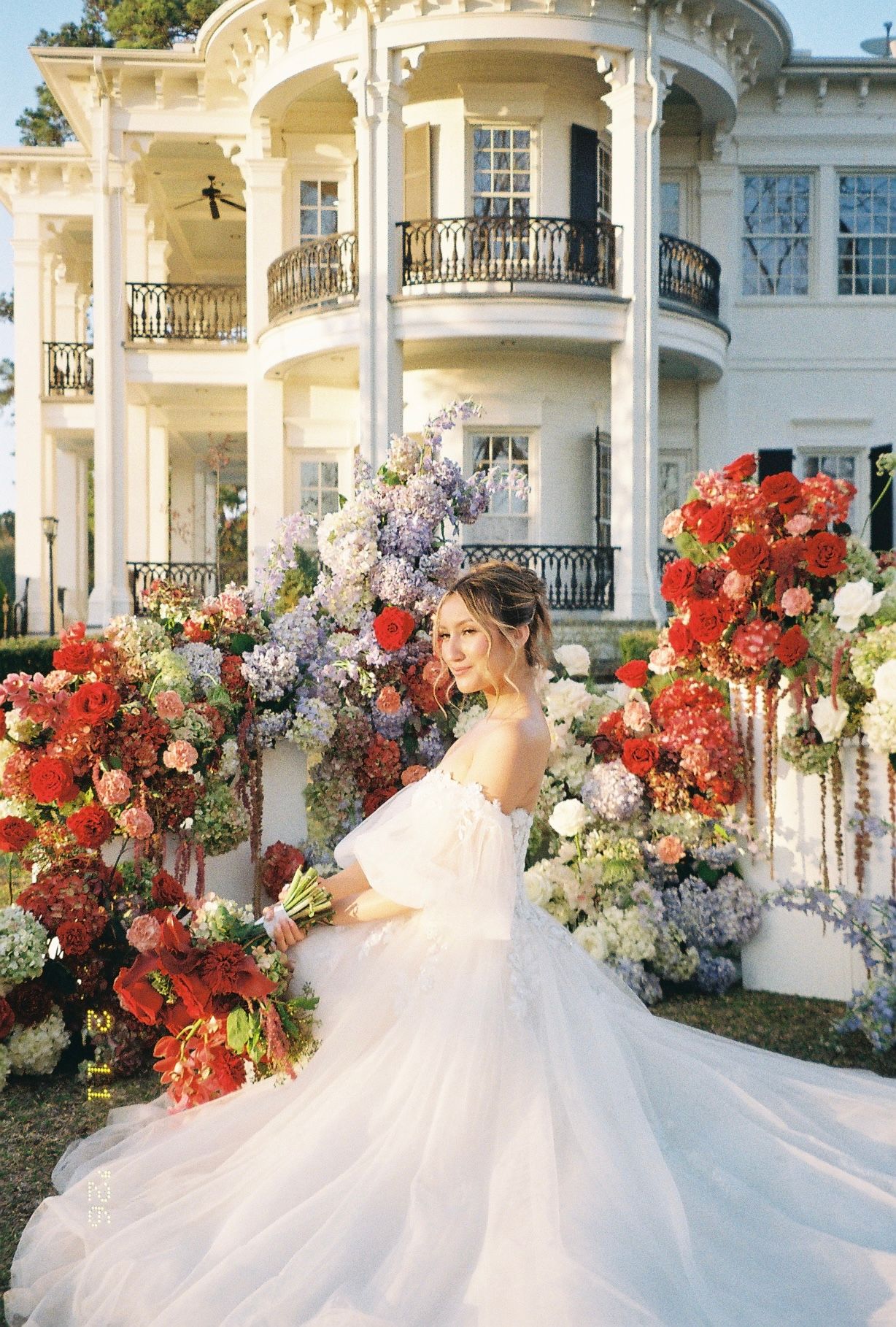 Bride in white gown surrounded by vibrant red and purple floral arrangements outside an elegant white mansion.