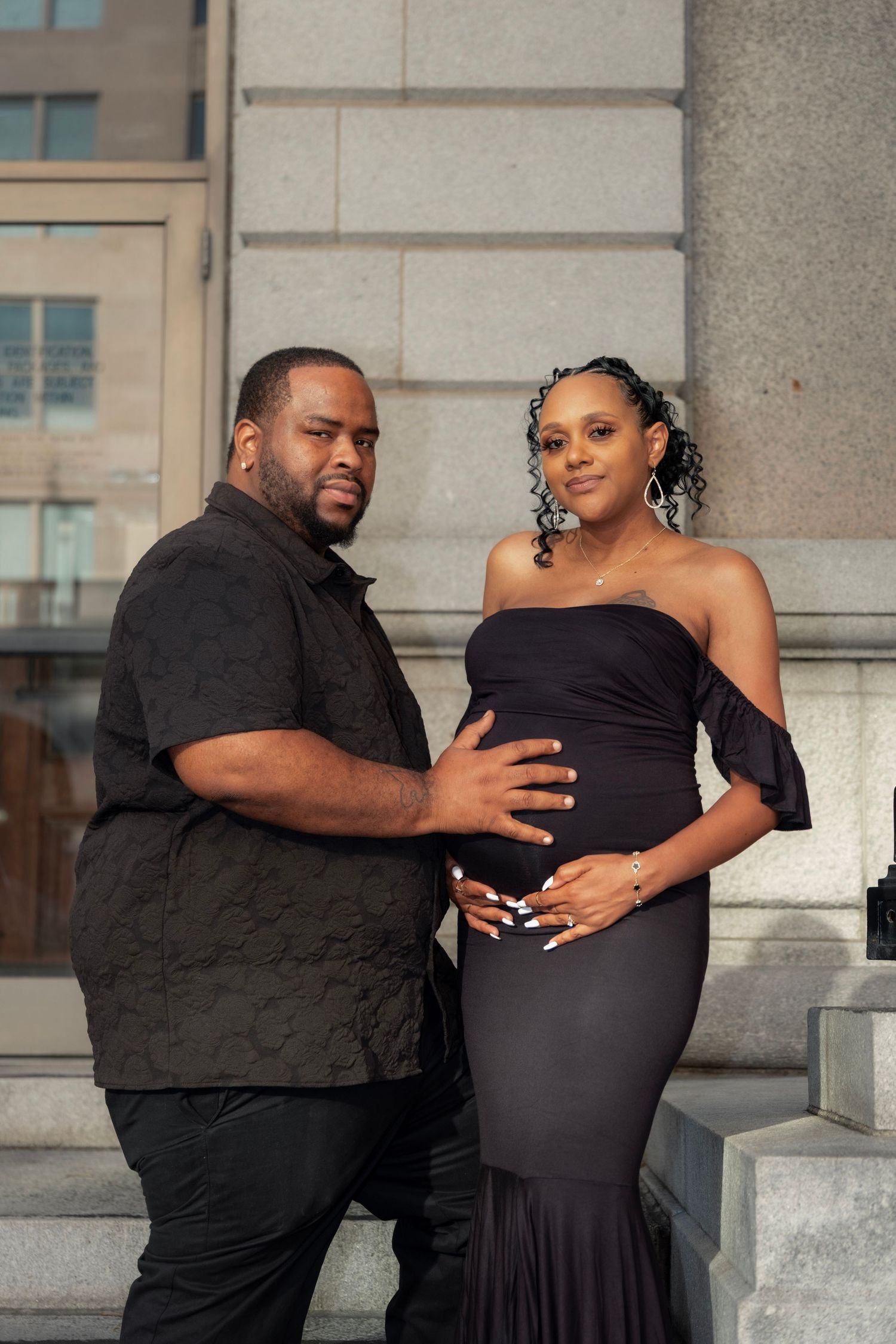 Couple in all-black outfits sharing a tender moment on steps, man gently touching pregnant woman's baby bump.