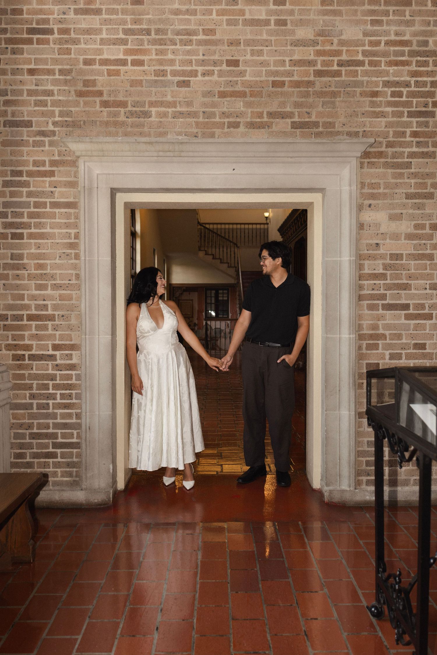 A couple holds hands in a brick archway doorway, woman in white dress and man in black shirt, with staircase behind them.