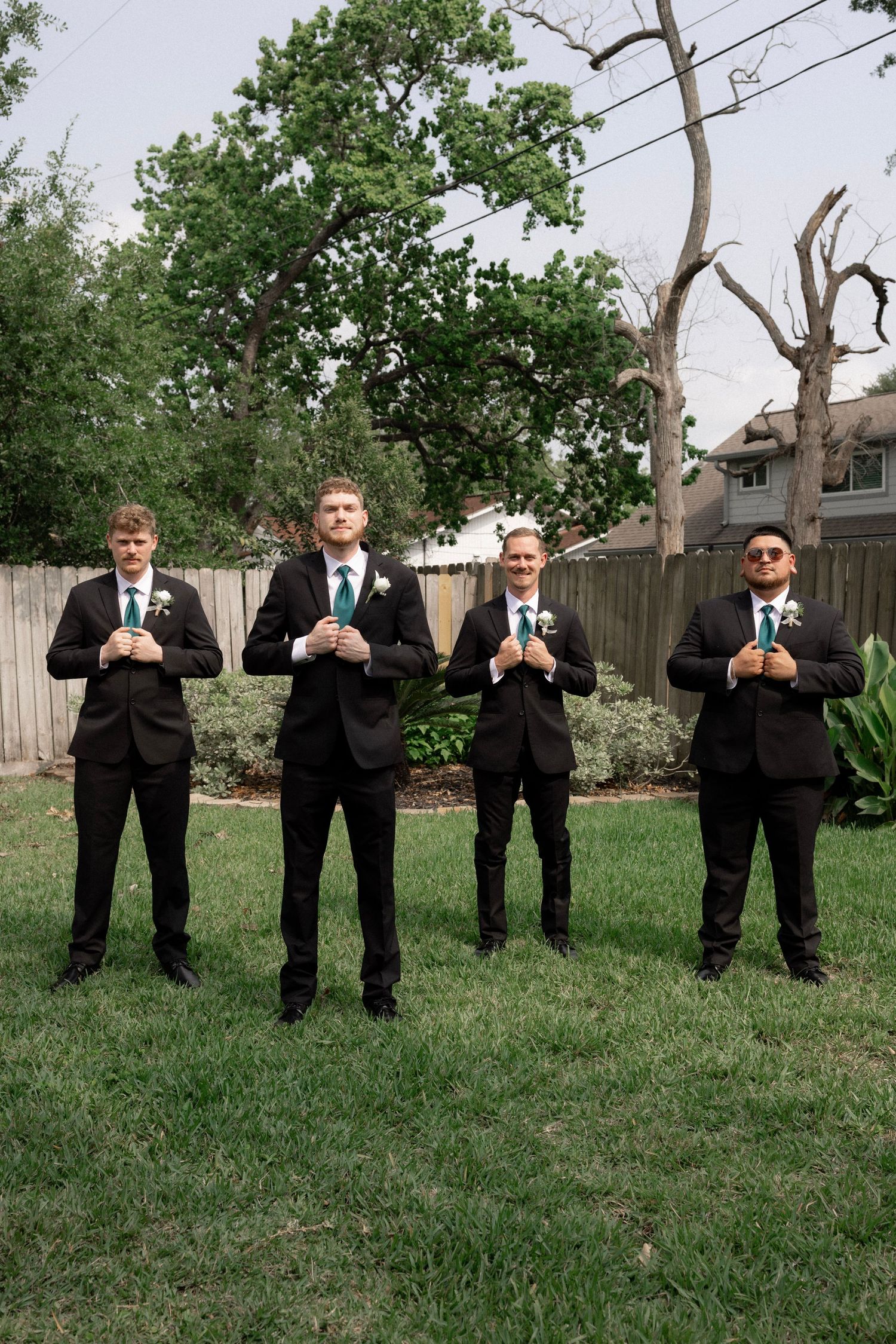 Four groomsmen in black suits with green ties pose confidently in a backyard.