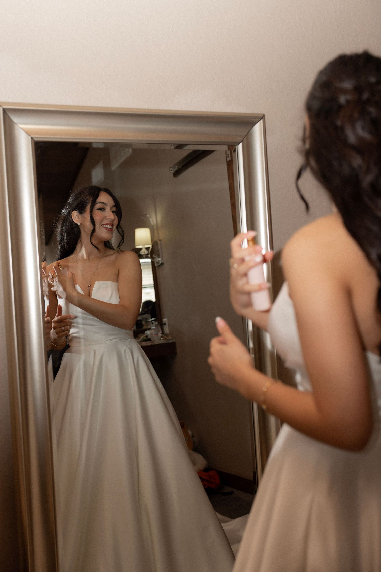 Smiling bride in strapless white ballgown admires her reflection in a large gold-framed mirror.