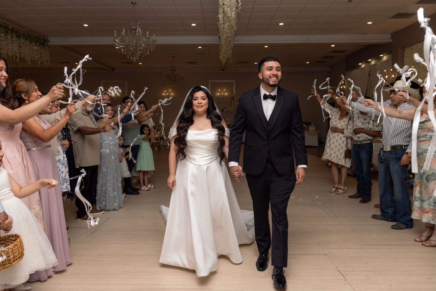 Newlyweds exit wedding reception through cheering guests holding white branches in elegant venue.