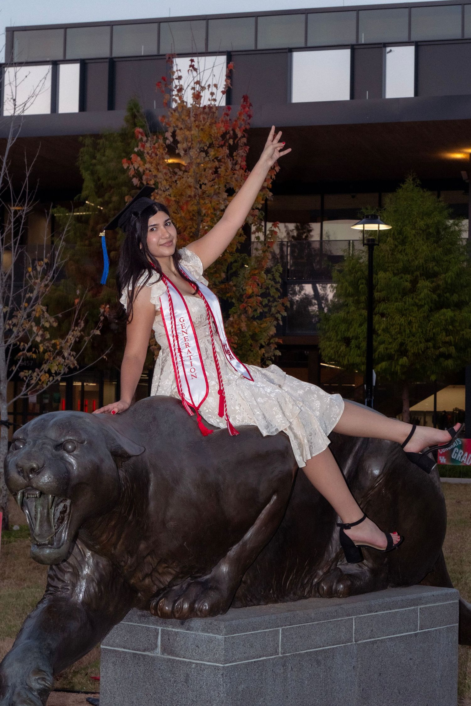 Graduate in white dress sitting on bronze cougar statue with arm raised at university campus.