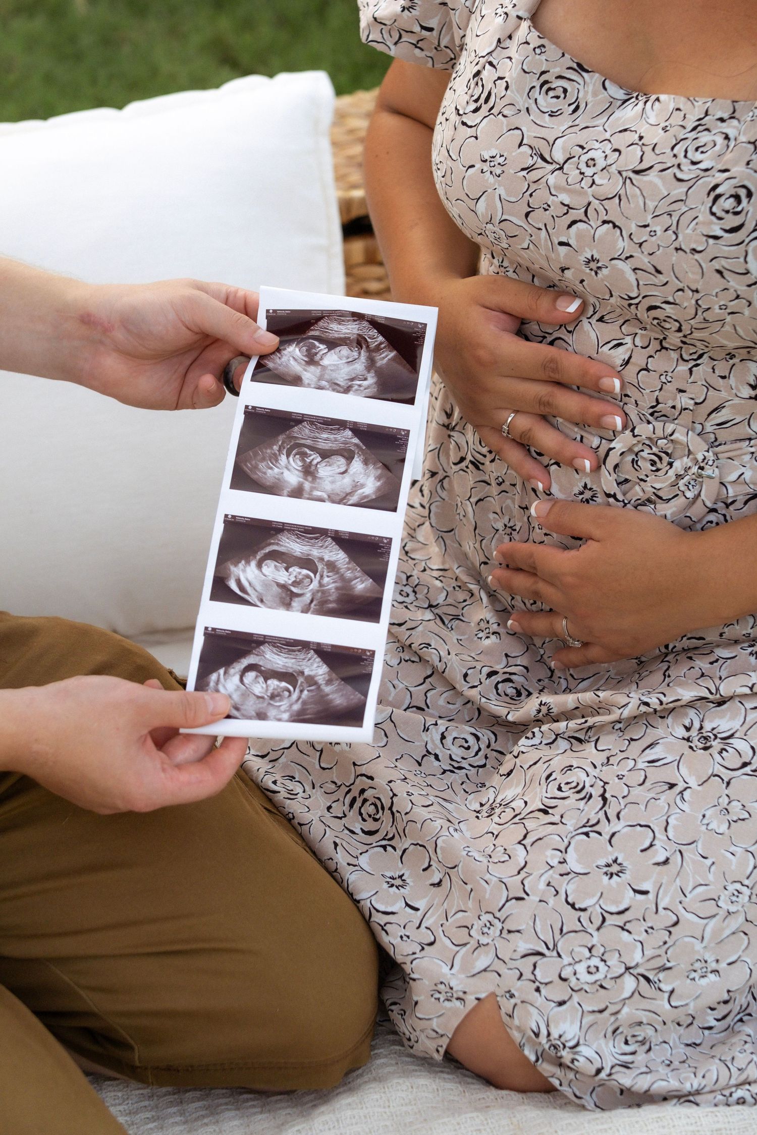 Pregnant woman in floral dress holds ultrasound photos while cradling baby bump outdoors.