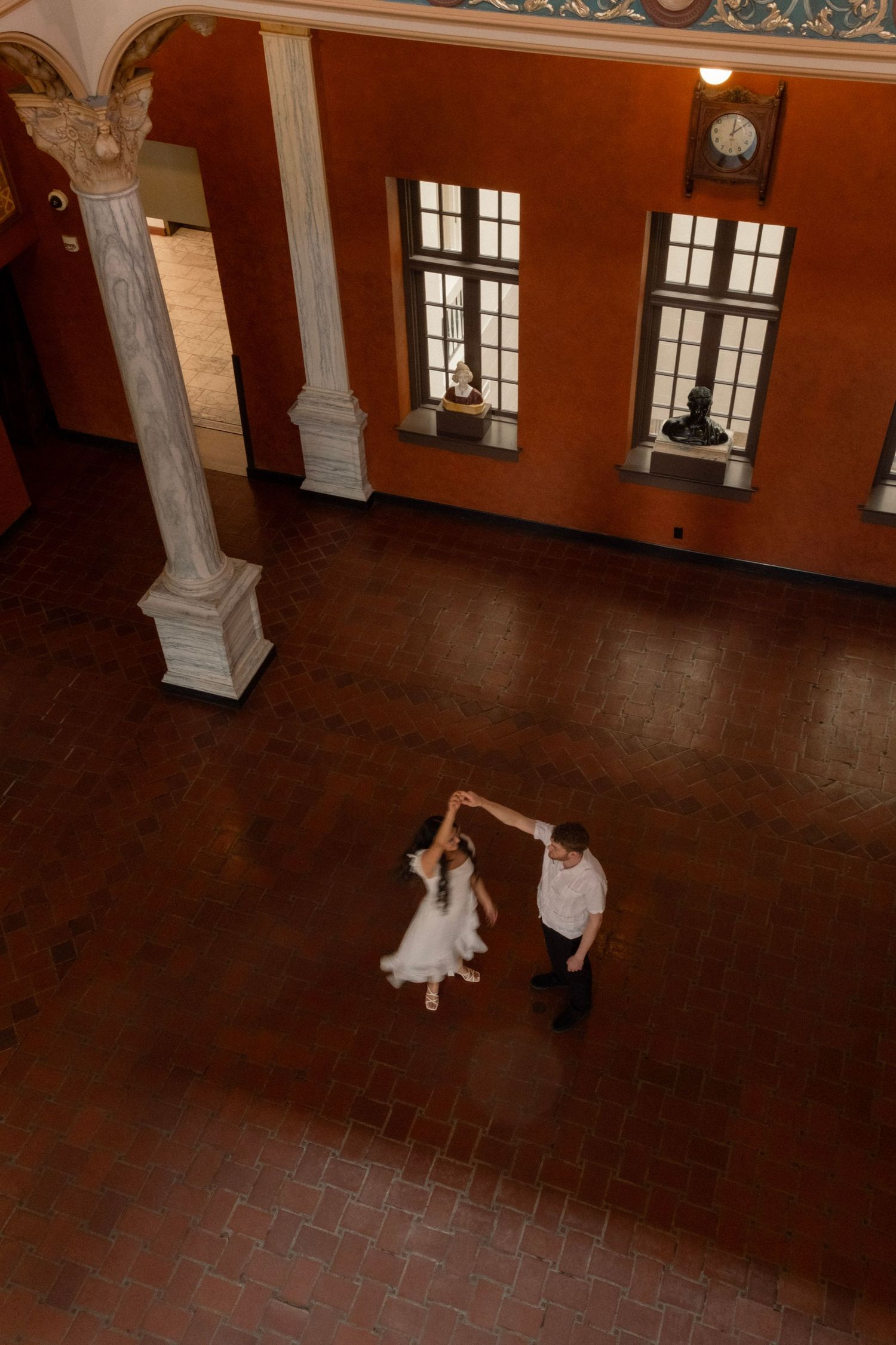 Aerial view of newlyweds sharing their first dance in a grand historic ballroom with tall columns.
