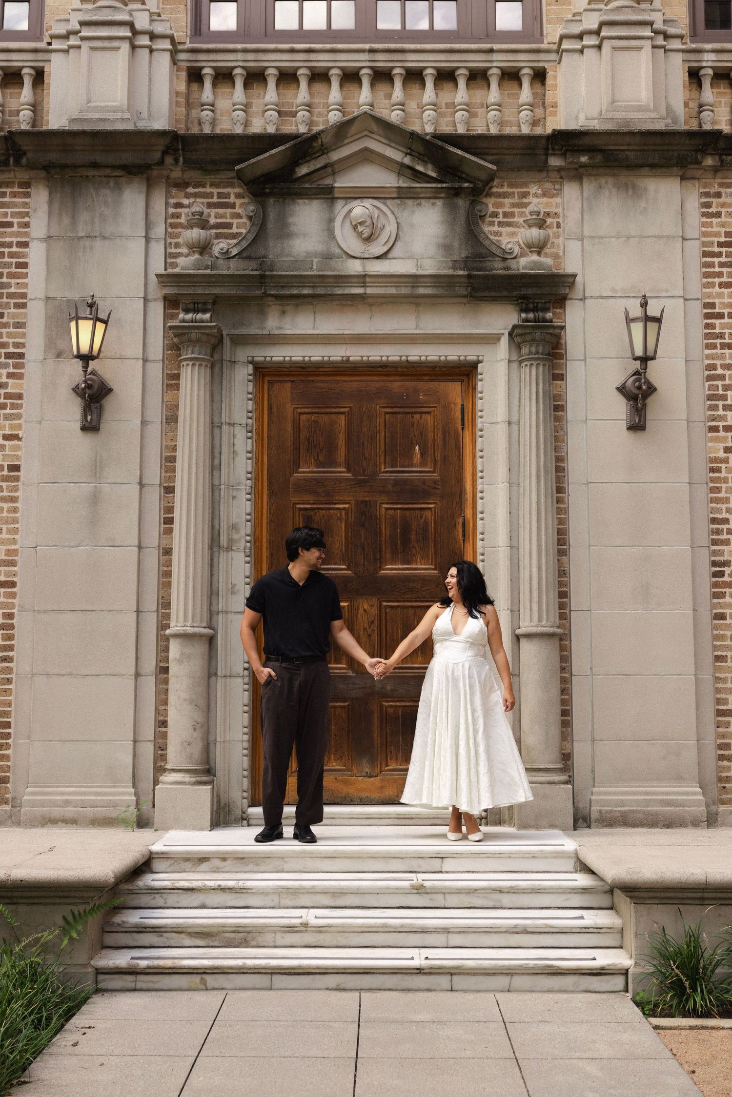 Couple holding hands on stone steps before ornate wooden door of historic brick building, woman in white dress, man in black.