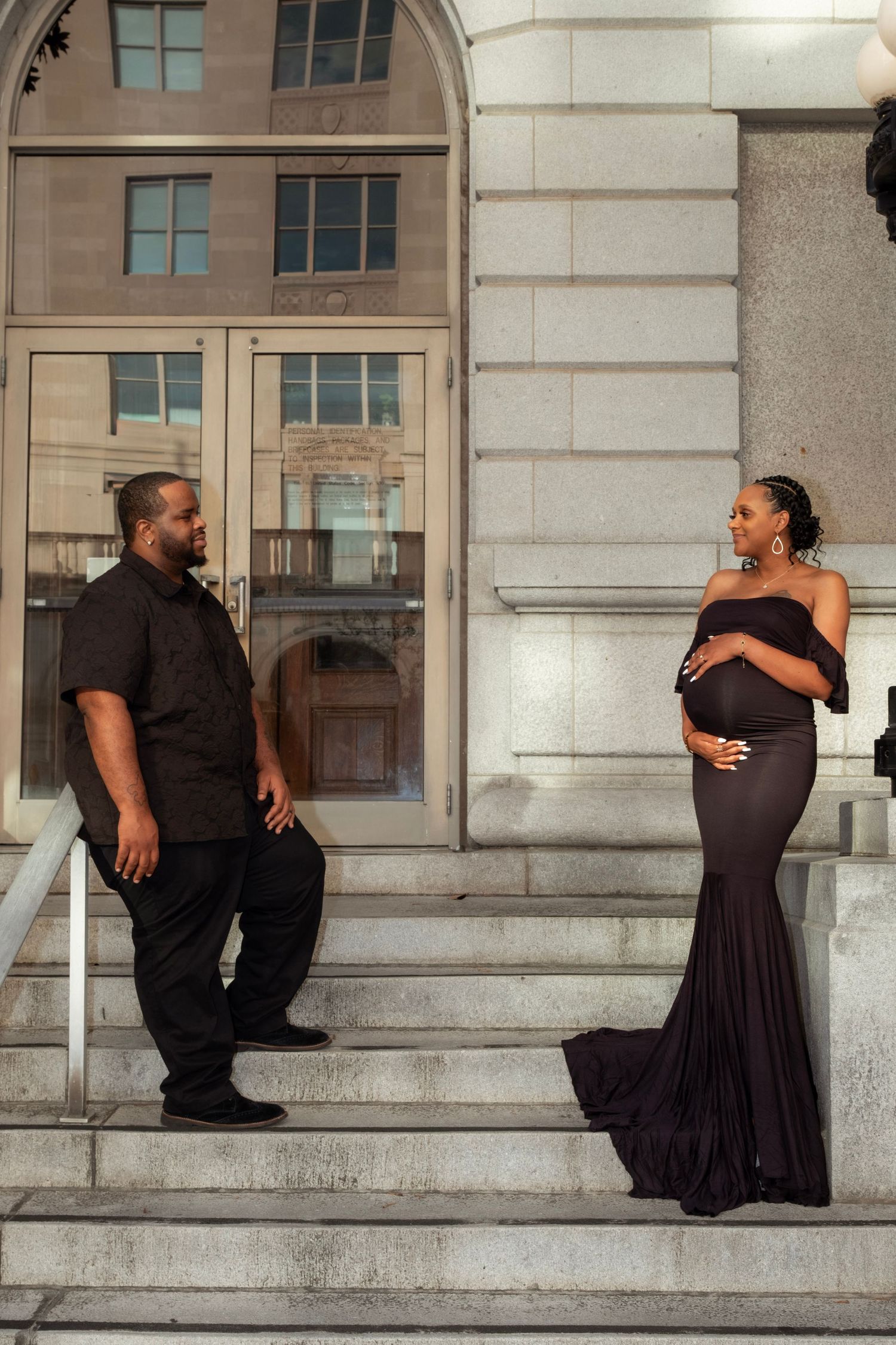 Expecting couple in matching black outfits posing on stone building steps during an urban maternity photo session.
