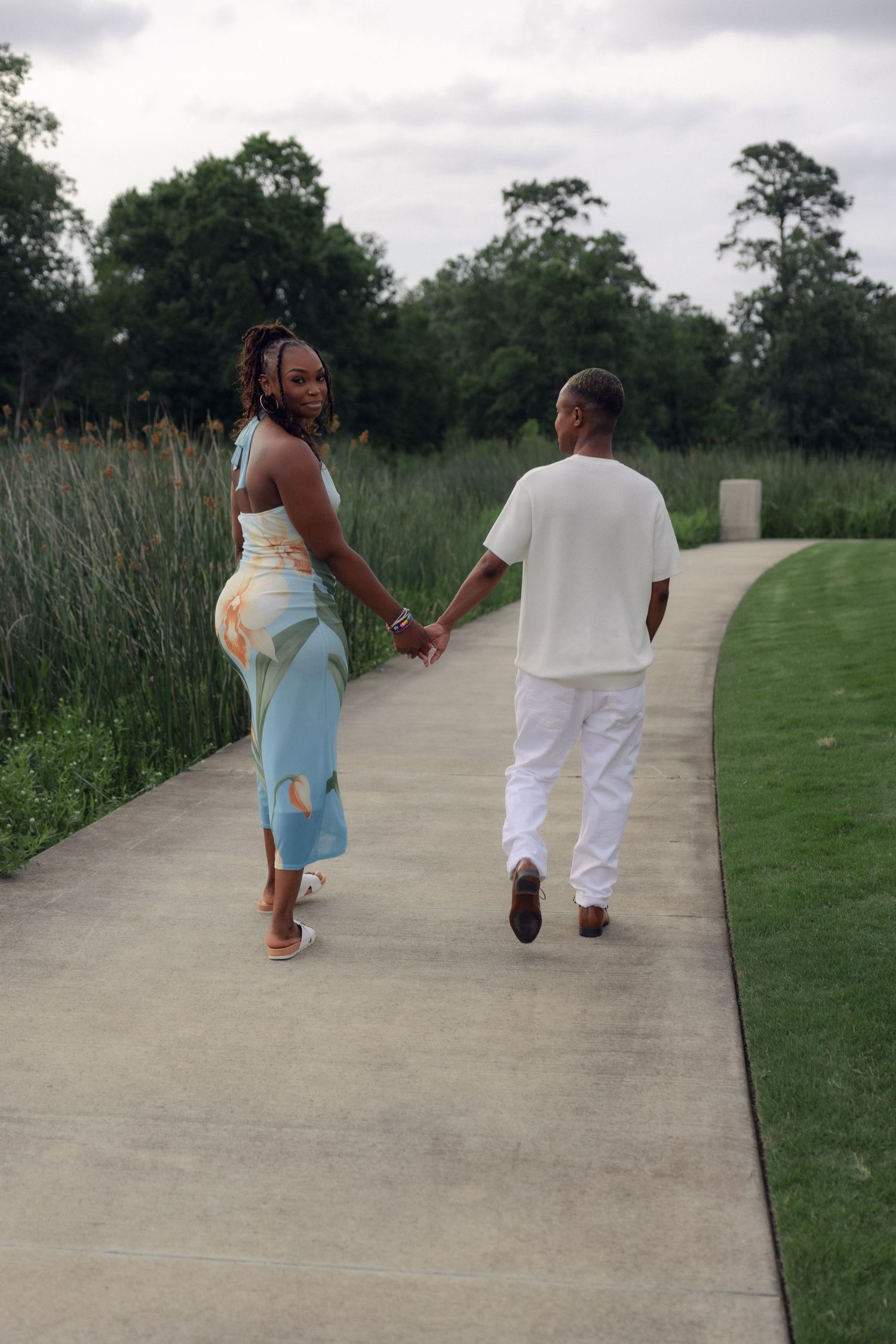 Couple holding hands walking along a curved pathway surrounded by tall grasses and green trees.