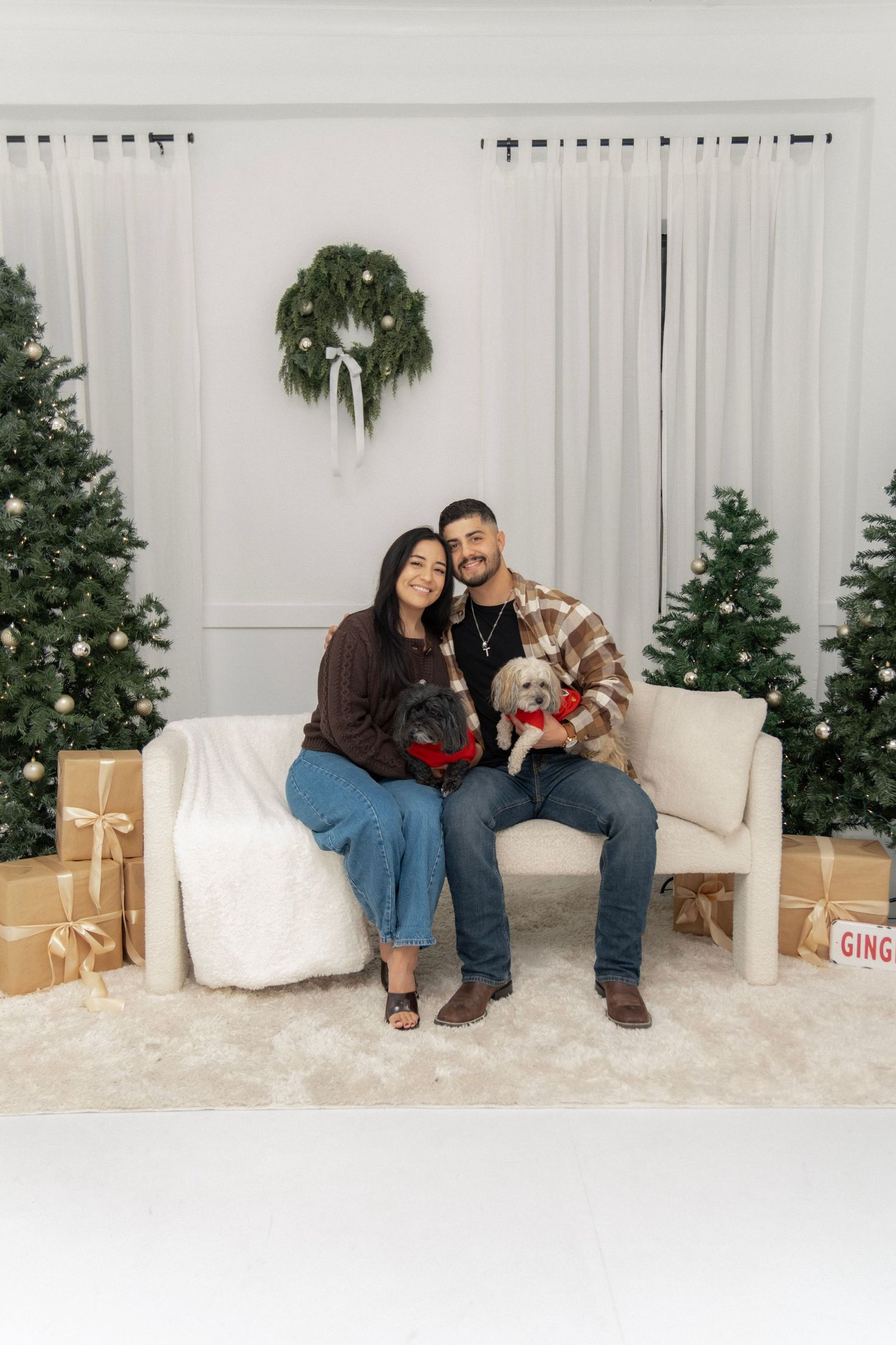 Couple sitting on white sofa with two small dogs in Christmas-themed photo with trees, gifts, and wreath backdrop.