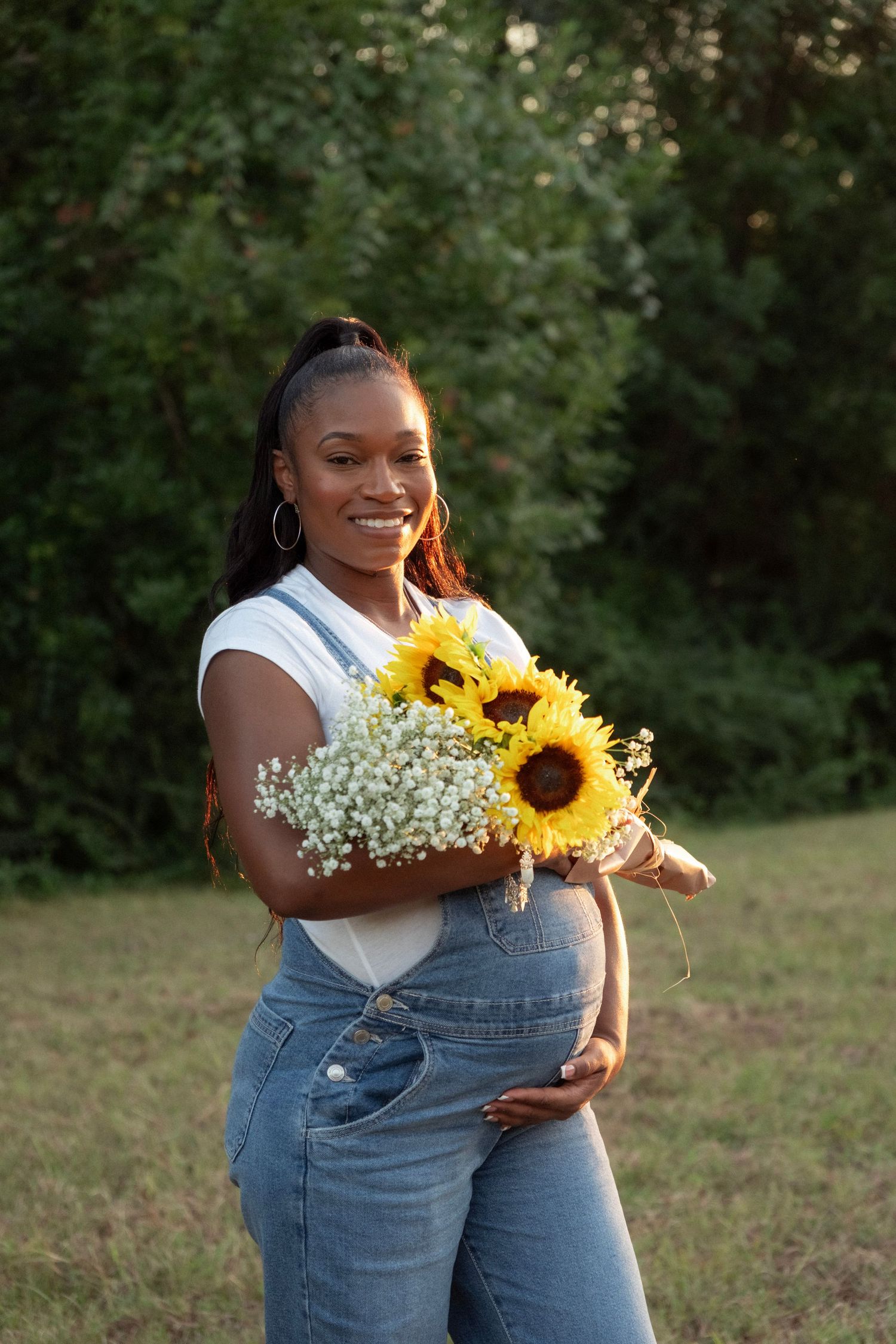 Smiling pregnant woman in denim overalls holding sunflowers and baby's breath bouquet during golden hour maternity shoot.