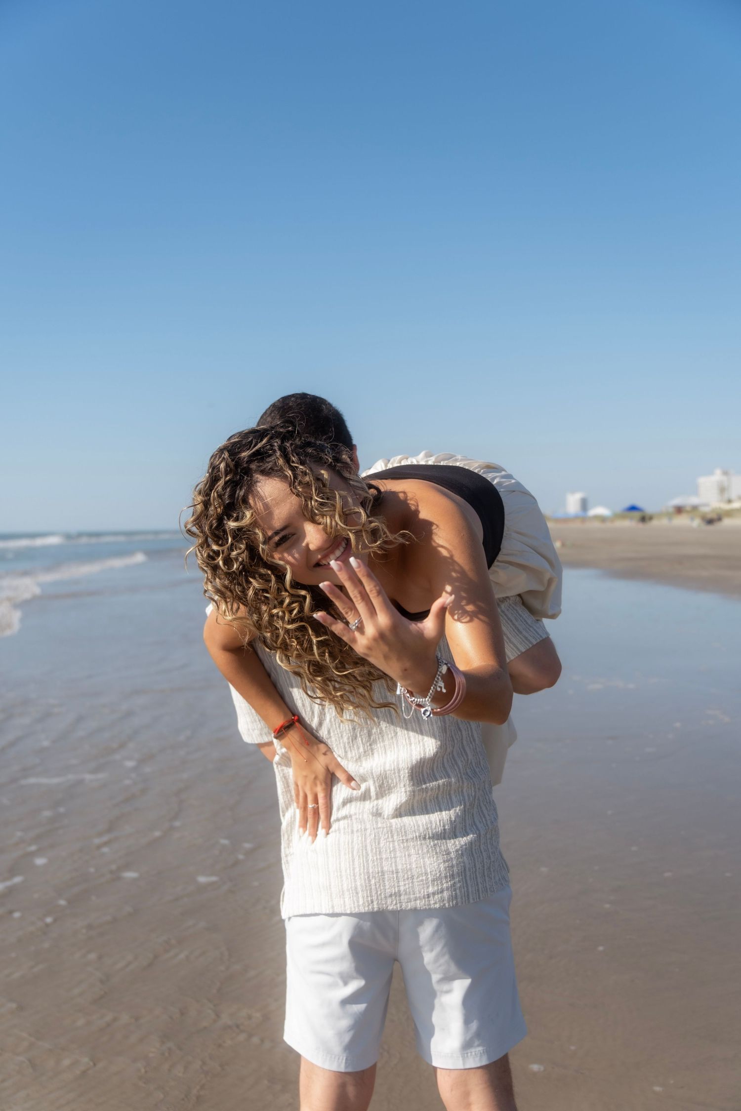 Couple embracing on sunny beach as woman shows engagement ring while laughing joyfully.