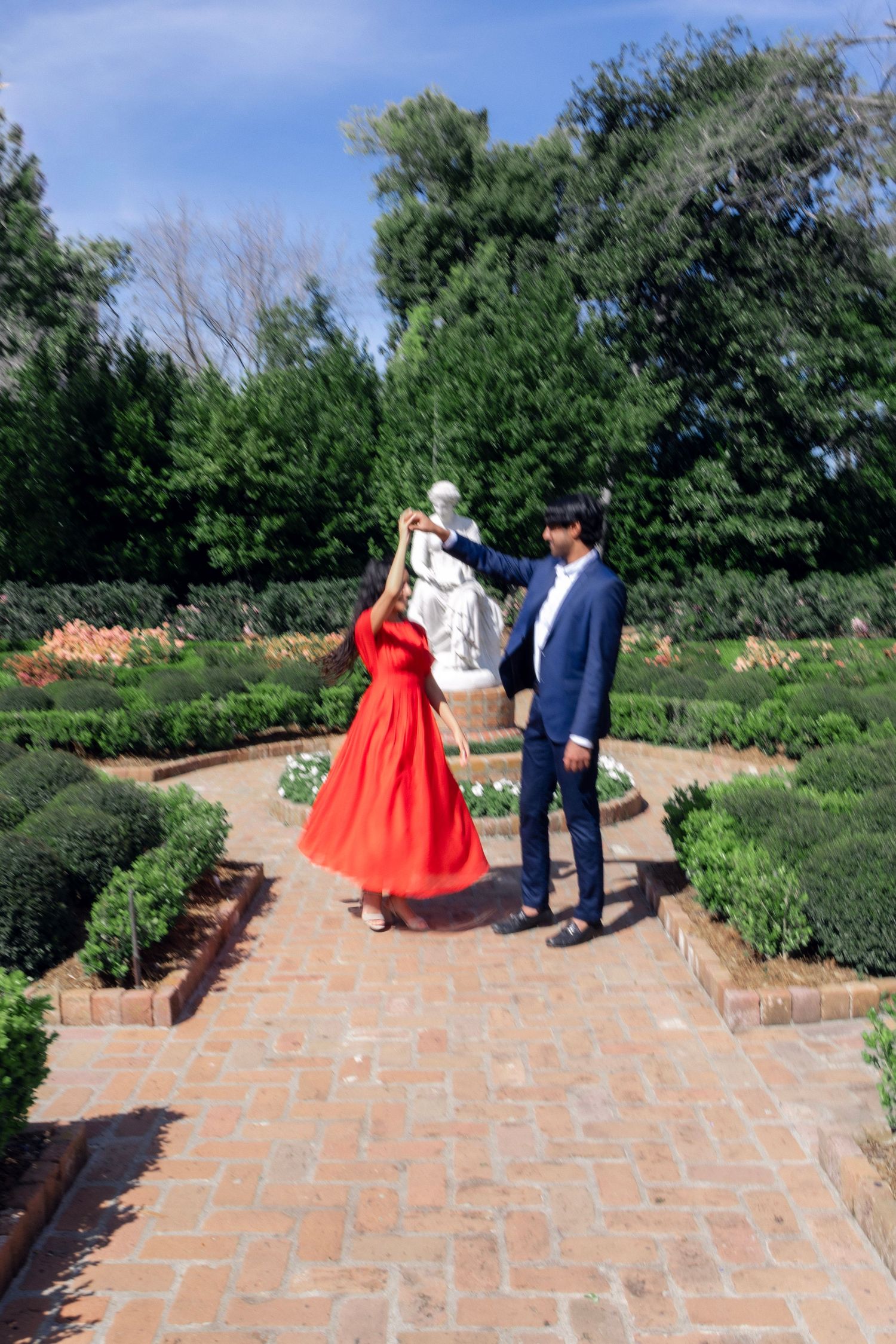 Couple dancing in a formal garden, woman in red dress spinning under man's hand in blue suit.