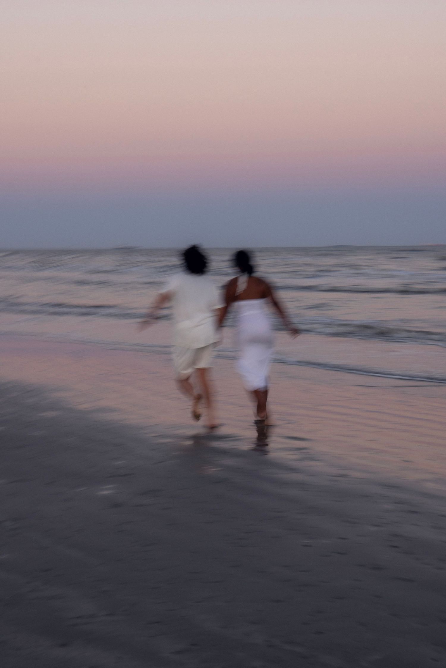 Blurred motion shot of couple in white running hand in hand on beach at sunset.