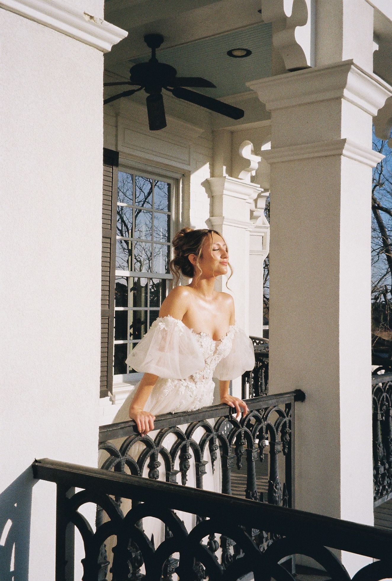 Elegant bride in off-shoulder white lace gown standing on ornate balcony of white mansion.
