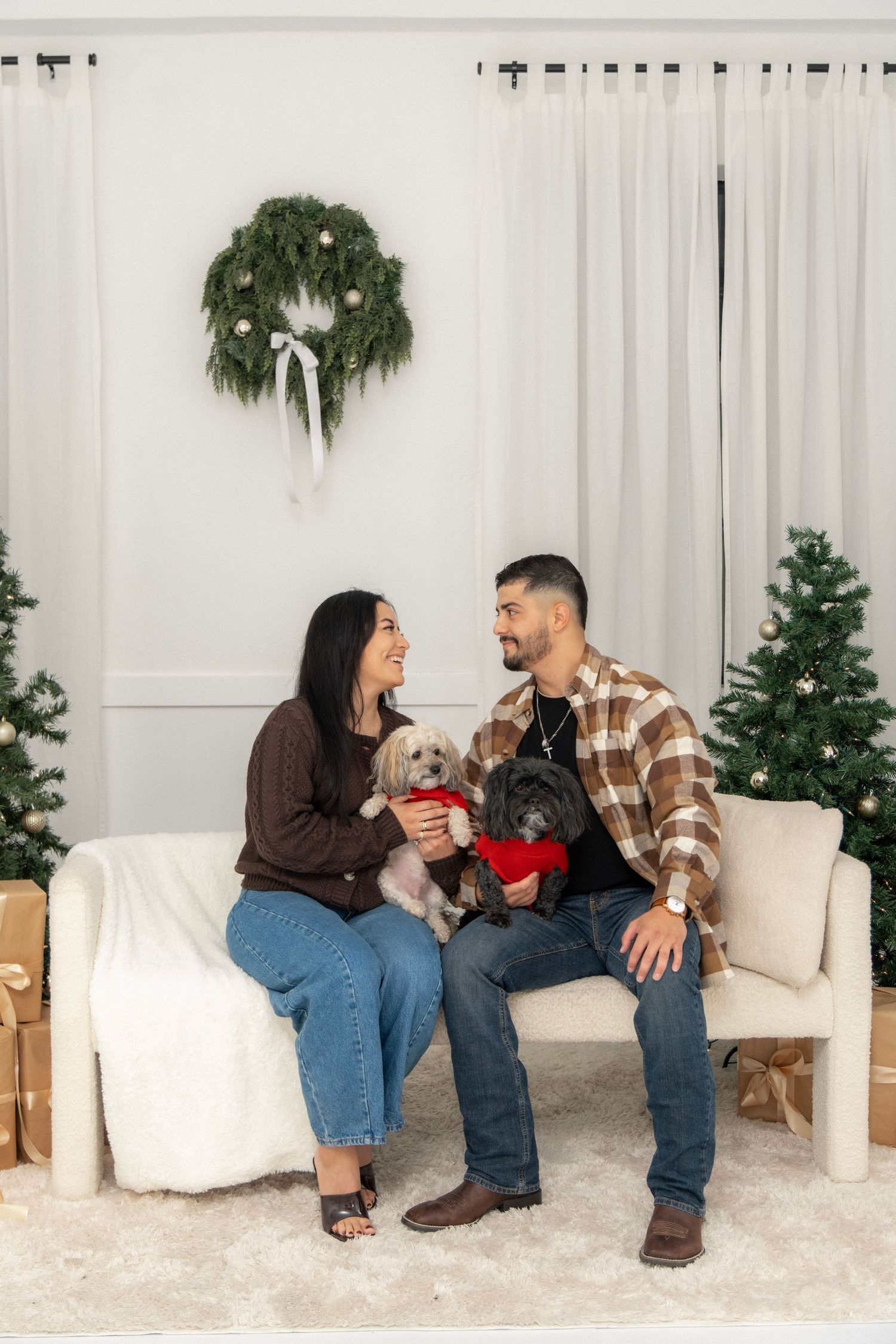 A couple sits on a white sofa with two small dogs amid Christmas trees and a wreath, sharing a joyful holiday moment.