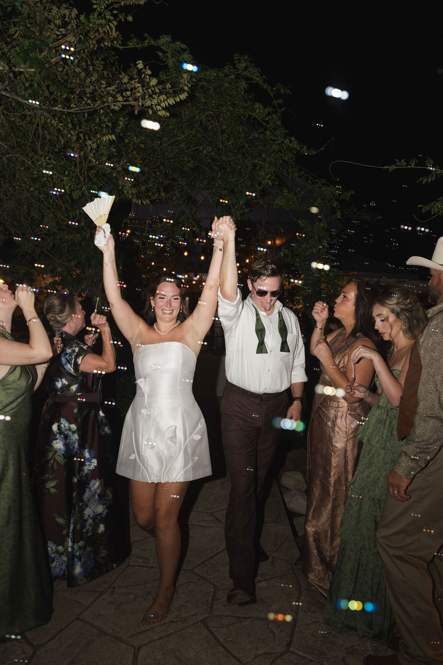 Bride in white mini dress and groom in sunglasses celebrate with raised hands at outdoor evening wedding reception.