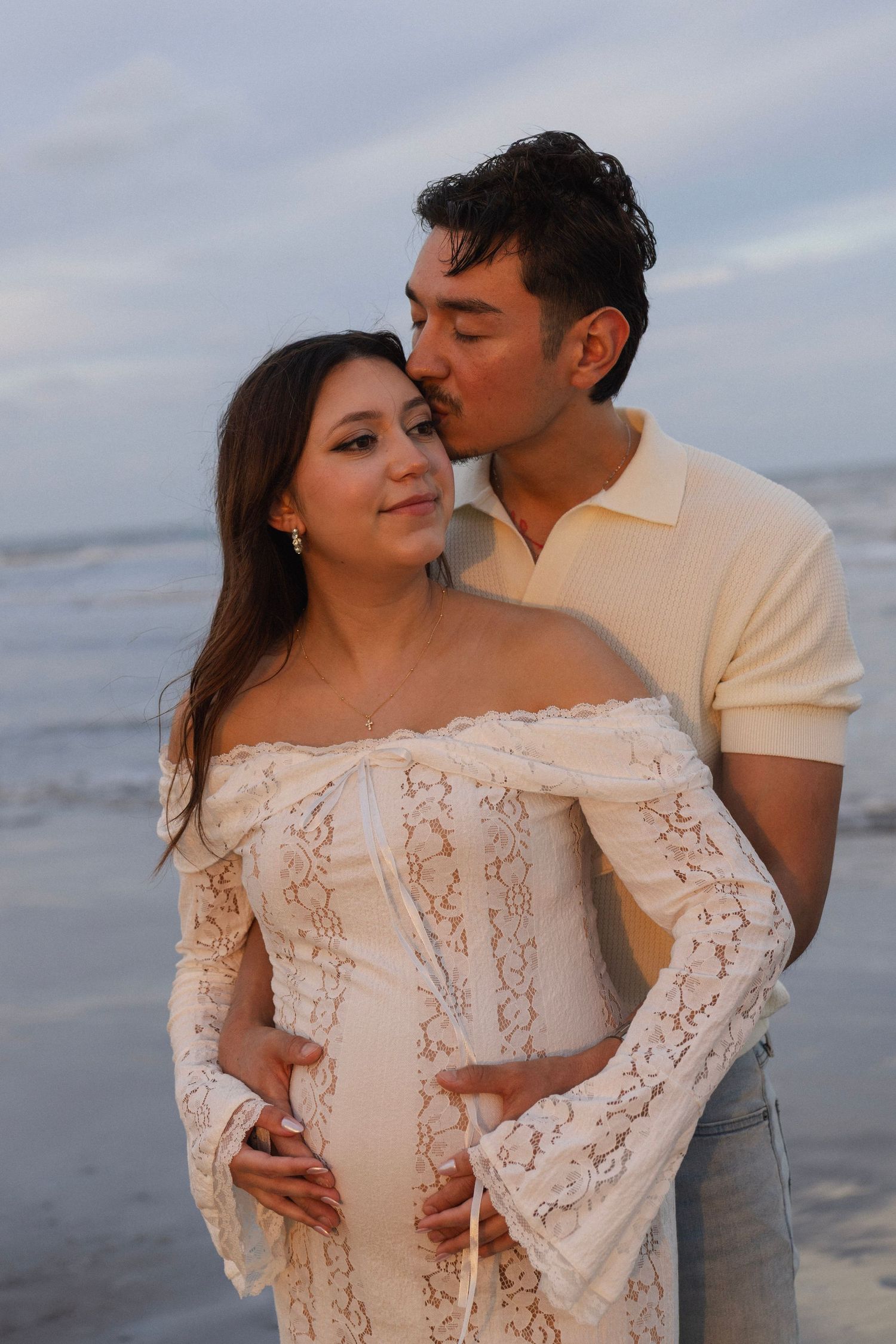 Couple sharing a tender moment on the beach, pregnant woman in white lace dress being embraced and kissed by partner.