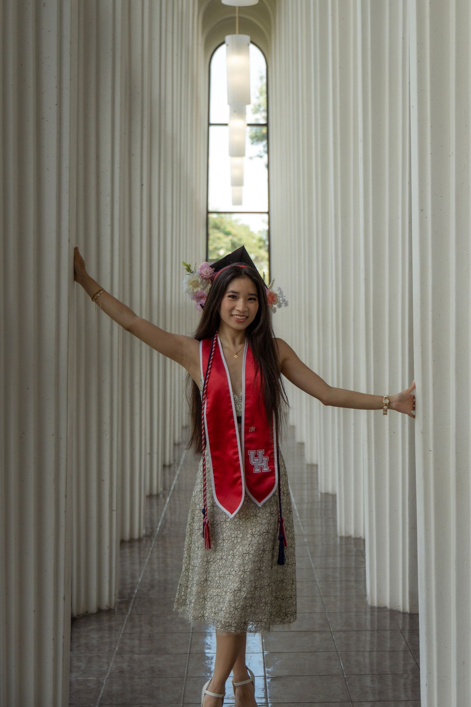 Graduate in UH red stole and cap with flowers posing between white columns with arms outstretched.