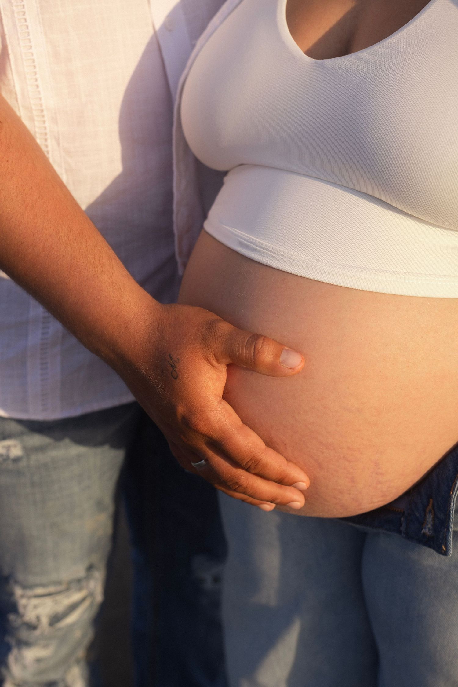 Close-up of a pregnant woman's bare belly being gently cradled by a partner's hand in warm sunlight.