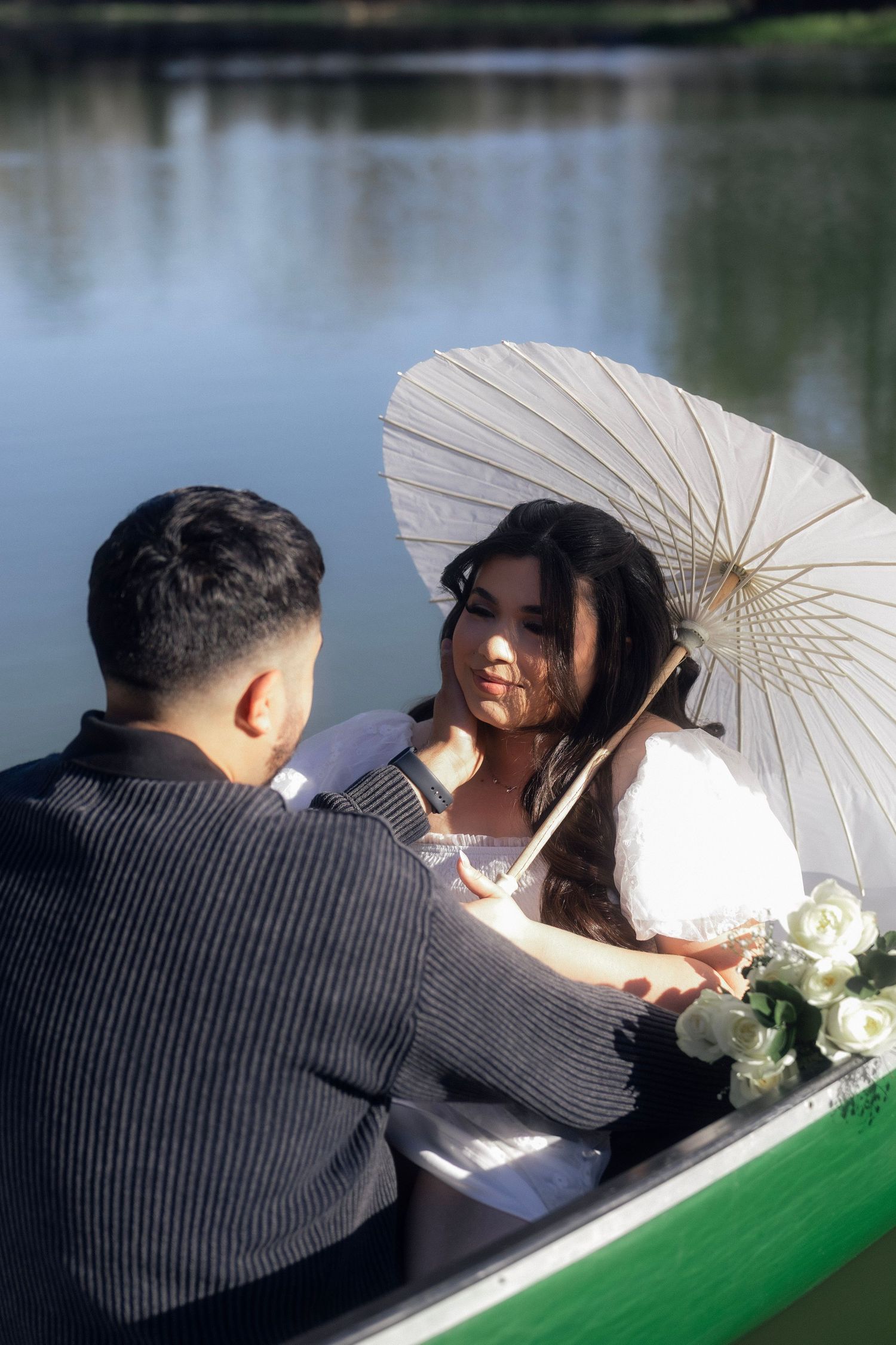 Romantic couple sharing intimate moment in a green rowboat on calm lake with white parasol.