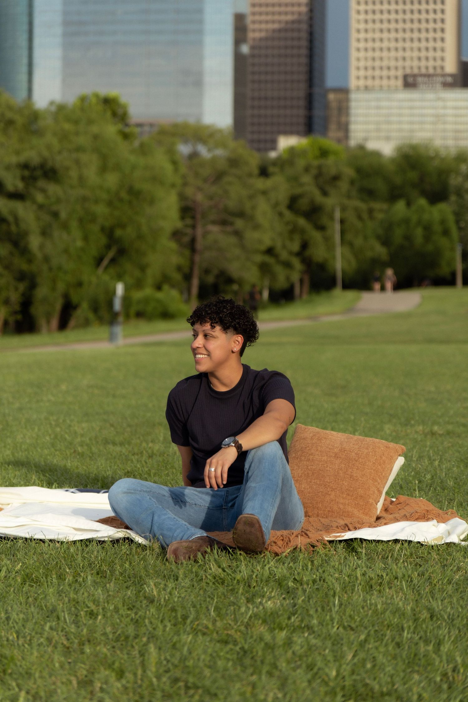 Smiling young man sits cross-legged on blanket in park with urban Houston skyline and trees in background.