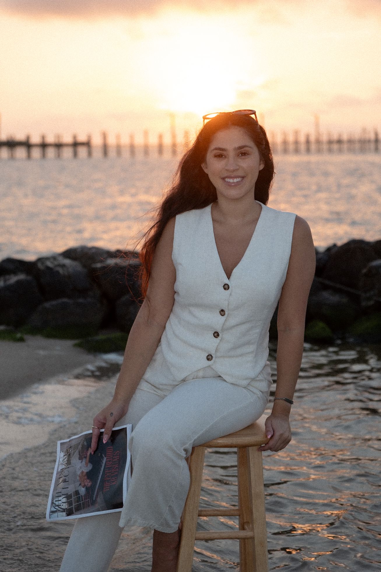Smiling woman in white linen outfit sits on wooden stool at sunset beach holding magazine.