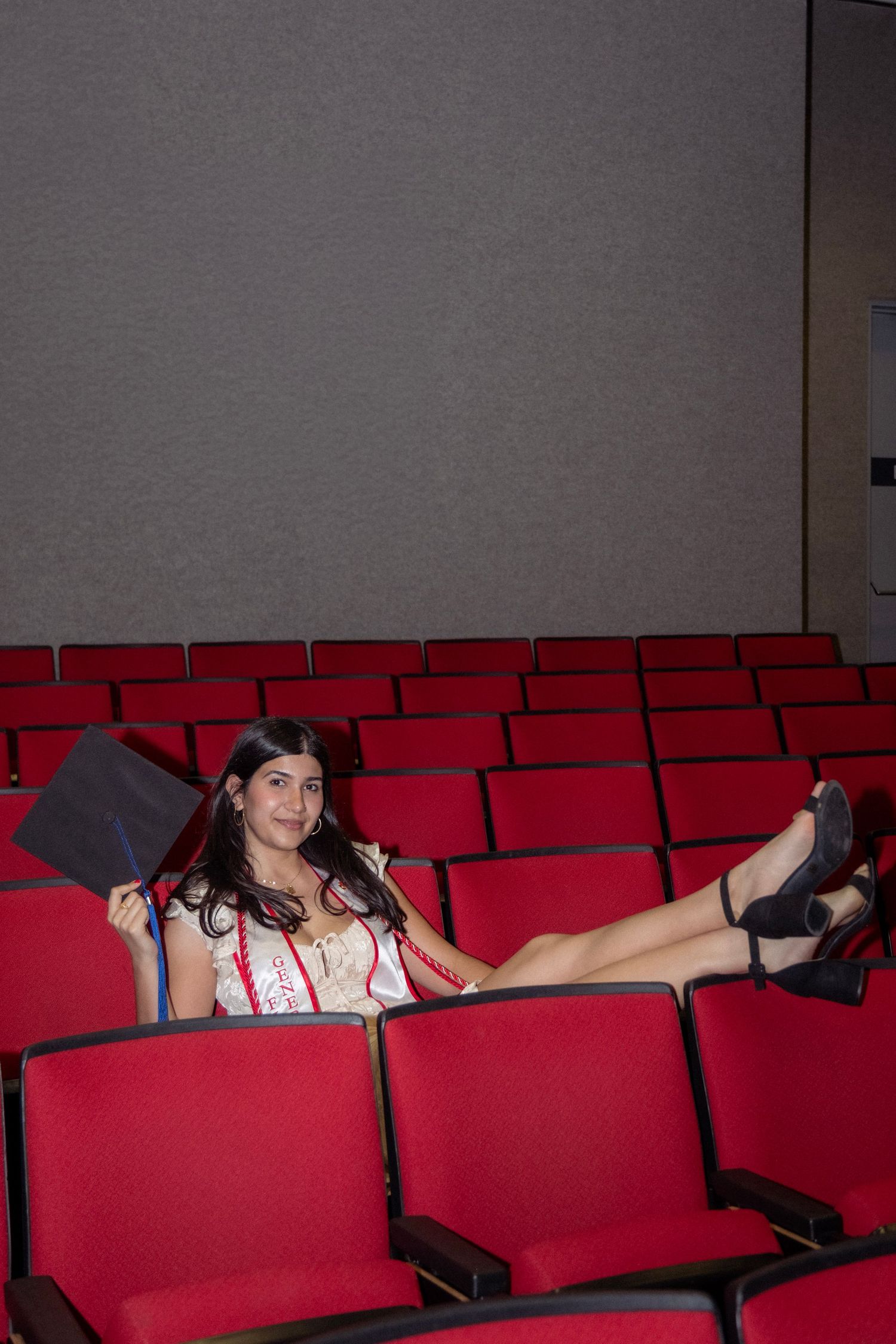 Female graduate relaxing in empty red auditorium seats holding graduation cap and wearing sash.