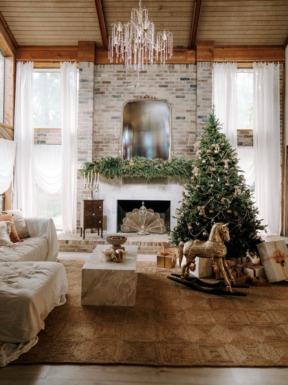 Elegant Christmas living room with decorated tree, crystal chandelier, brick fireplace, garland mantel, and golden rocking horse.