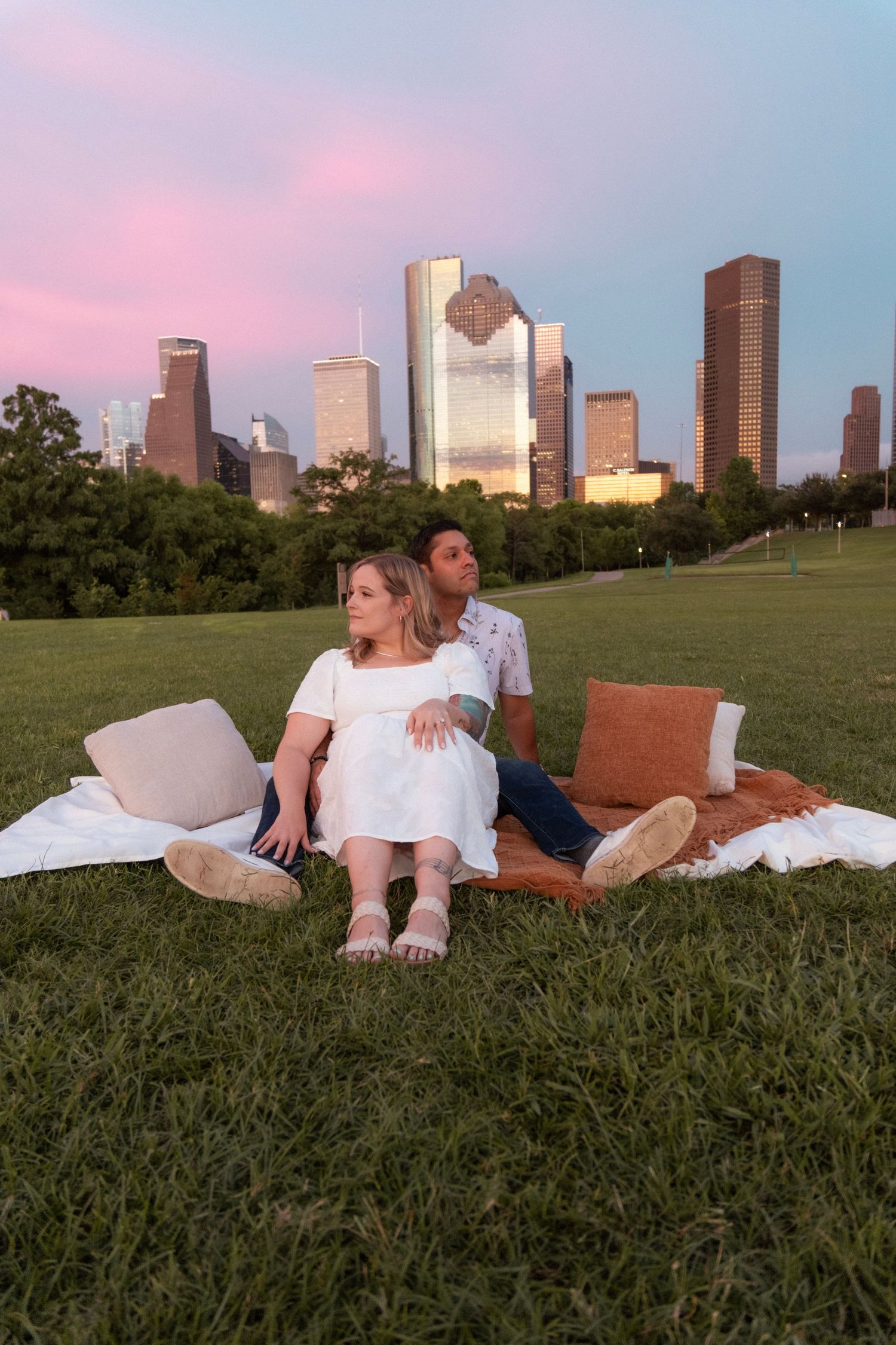 Couple sitting on a blanket in a park with the Houston skyline and pink sunset sky in the background.