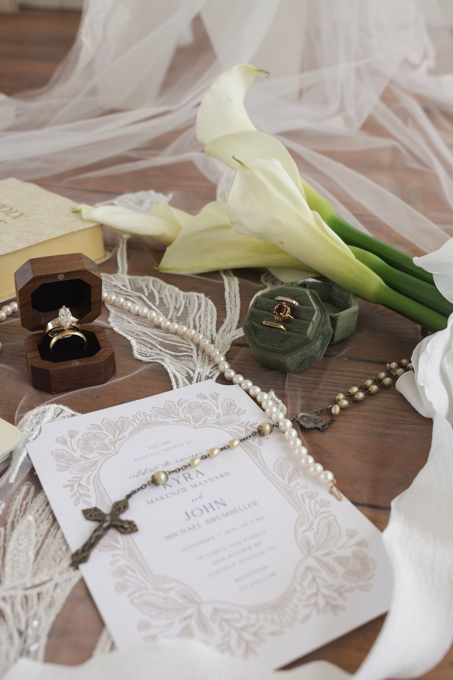 Elegant wedding flat lay with gold rings, pearl rosary, calla lilies, velvet ring boxes, and floral invitation on wood.