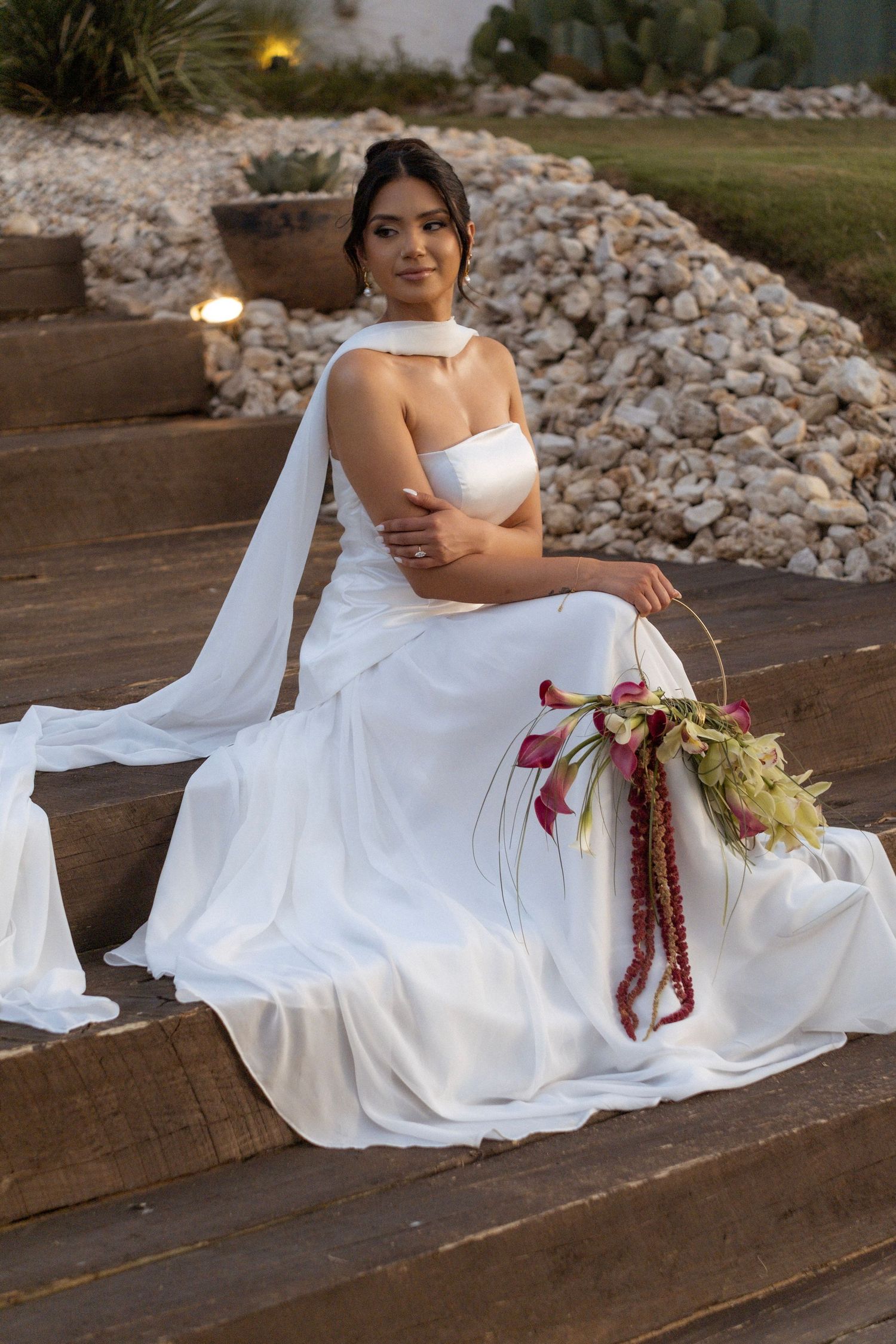 Bride in white gown with veil sits on wooden steps holding a cascading bouquet with red accents at an outdoor venue.