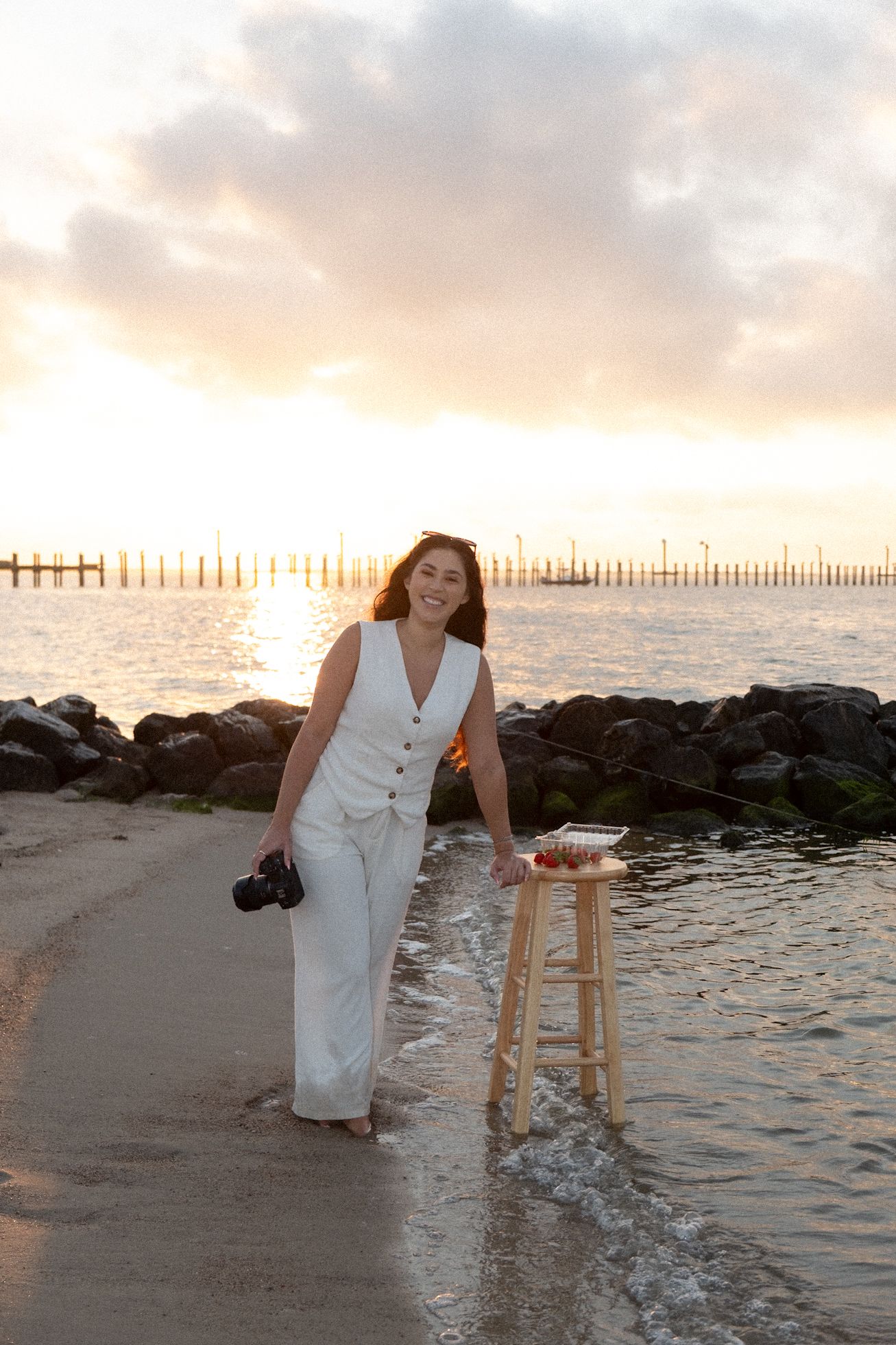 Female photographer in white outfit holds camera on rocky beach at golden sunset hour.