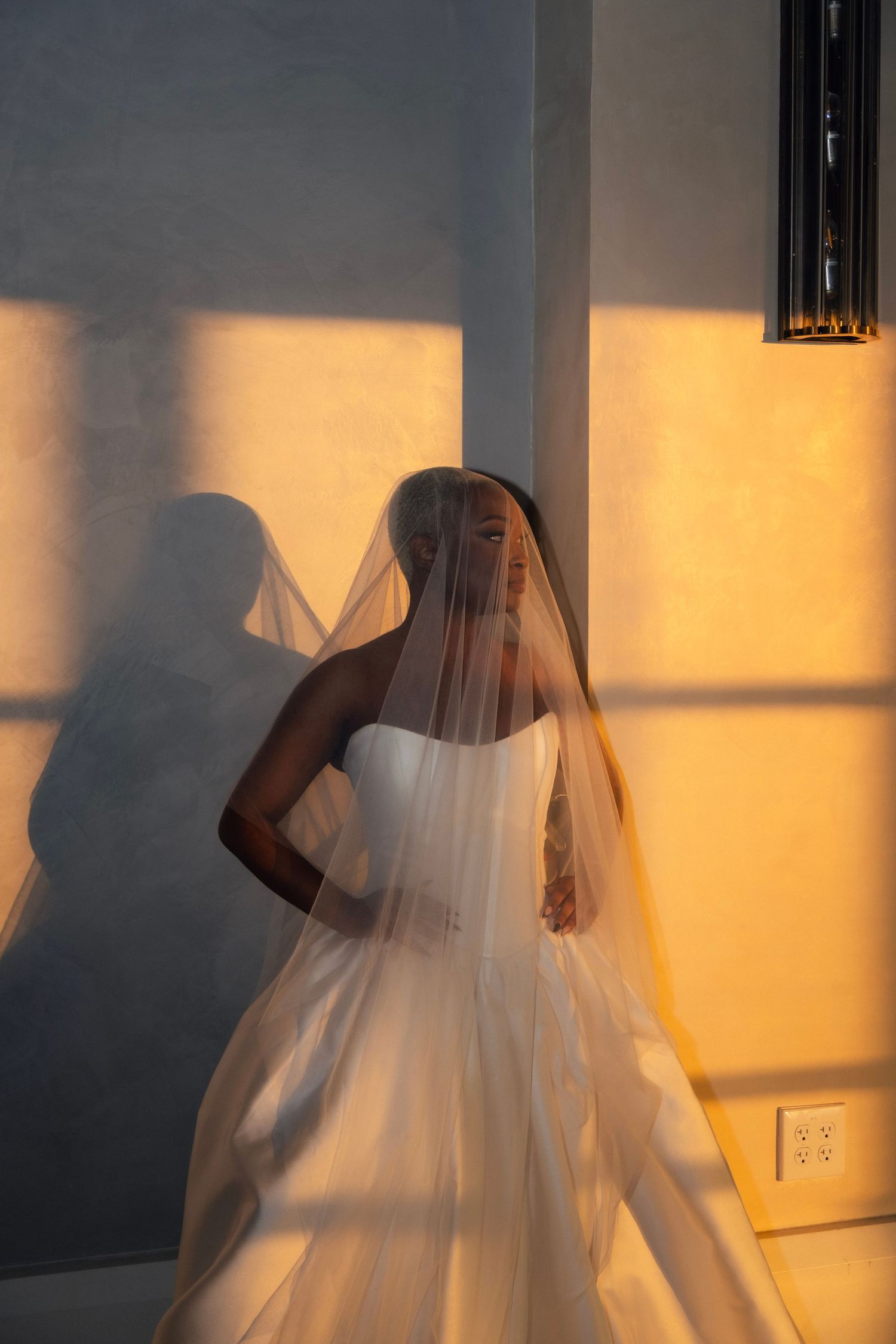 Bride in white strapless ball gown and cathedral veil bathed in warm golden sunlight against a gray wall.