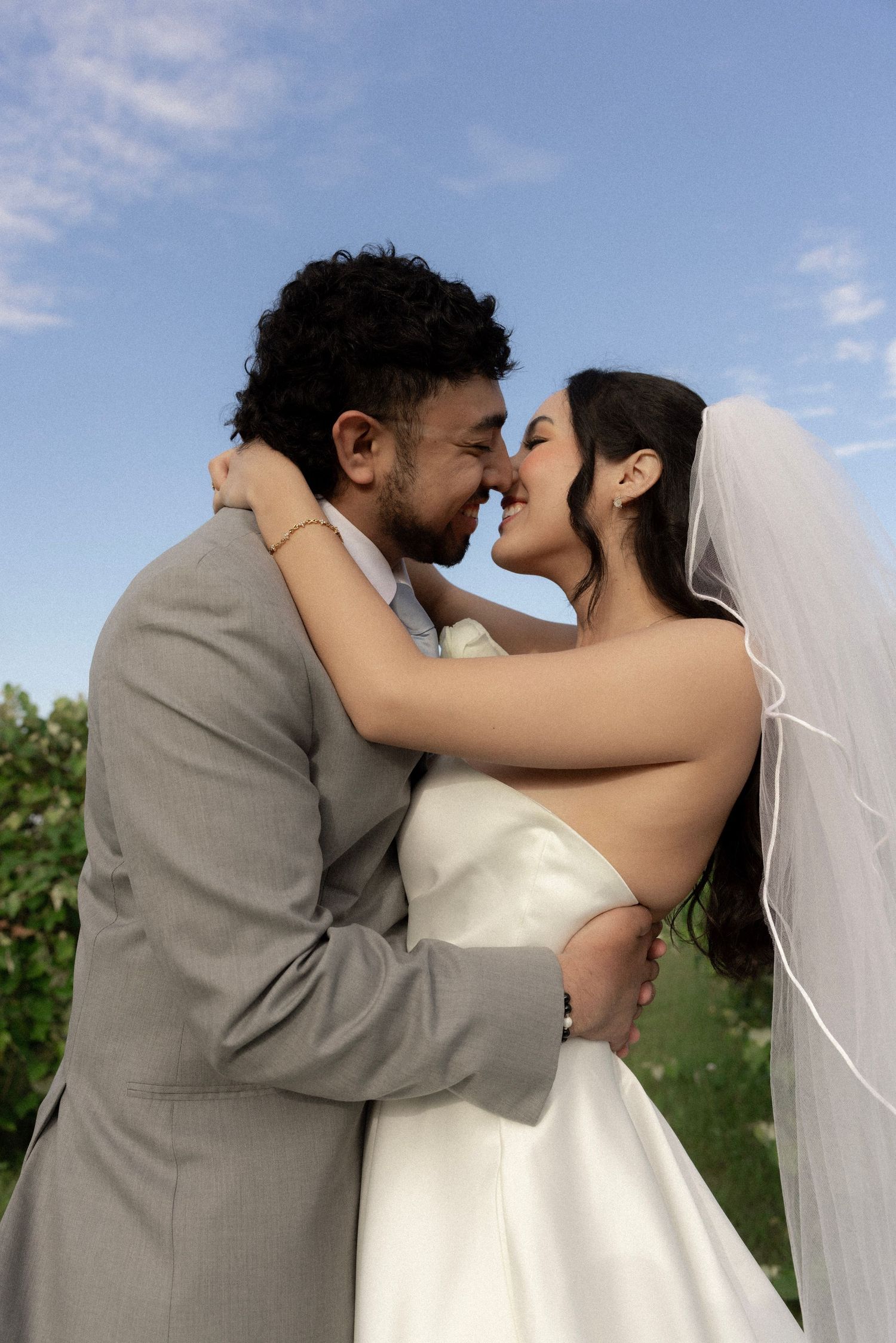 Smiling bride and groom embrace closely, touching foreheads in a tender moment at outdoor wedding.