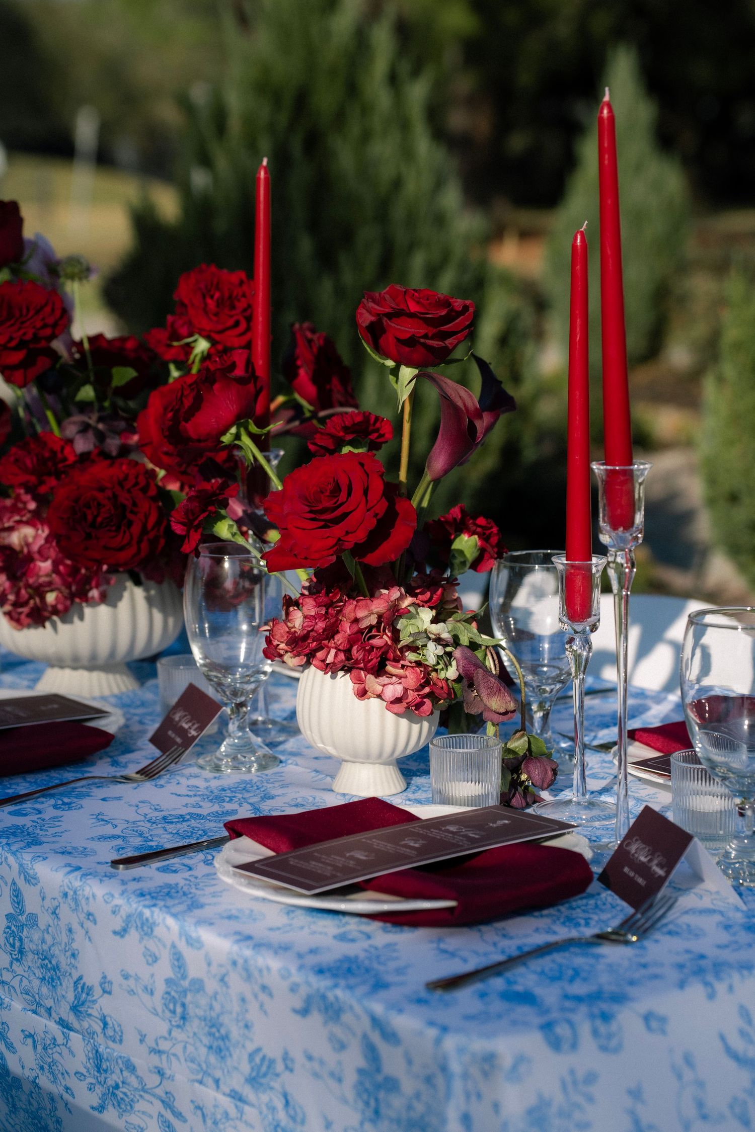 Elegant wedding tablescape with red roses, tall red taper candles, and crystal glassware on a blue toile tablecloth.