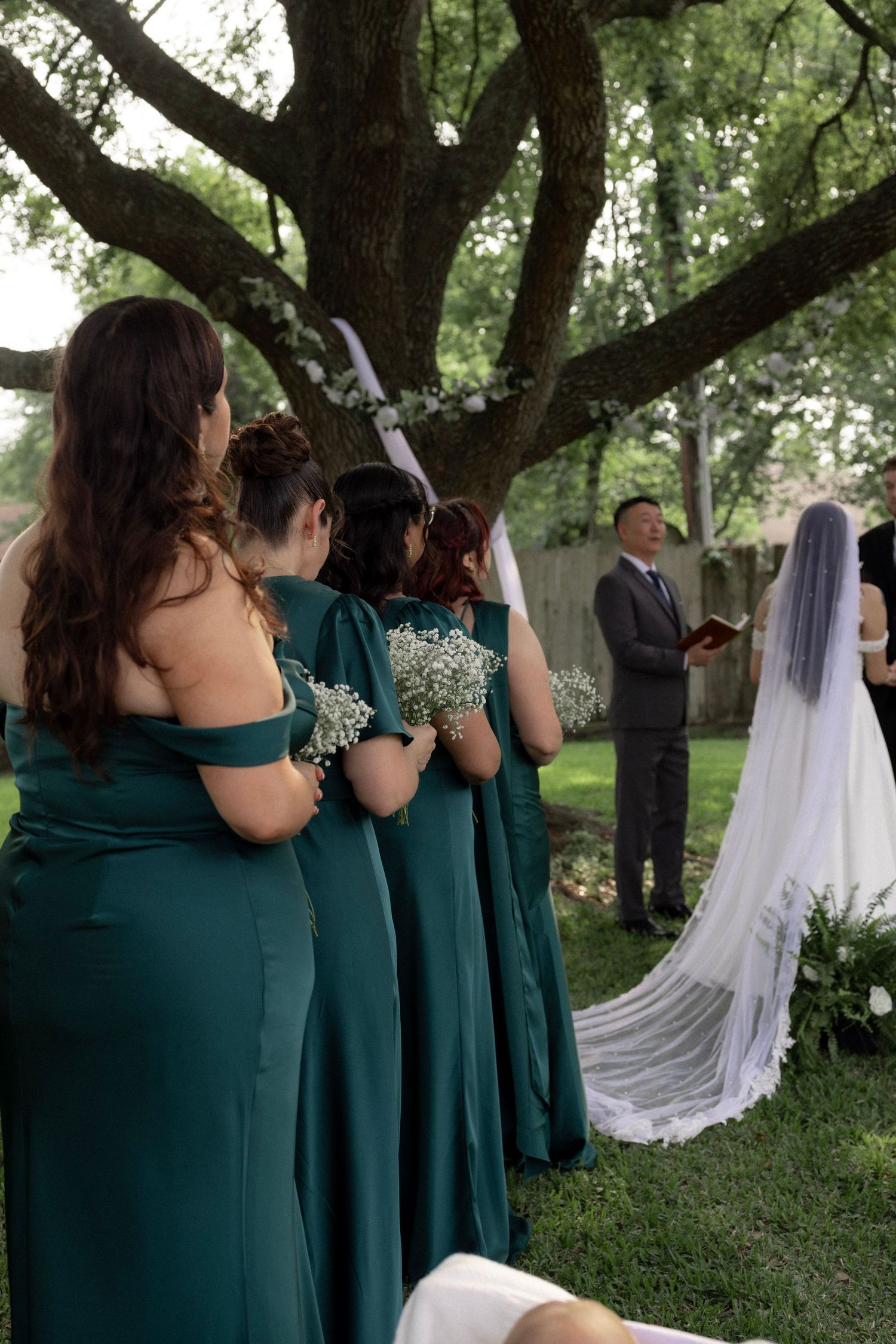 Bridesmaids in emerald gowns holding bouquets stand during an outdoor backyard wedding ceremony.