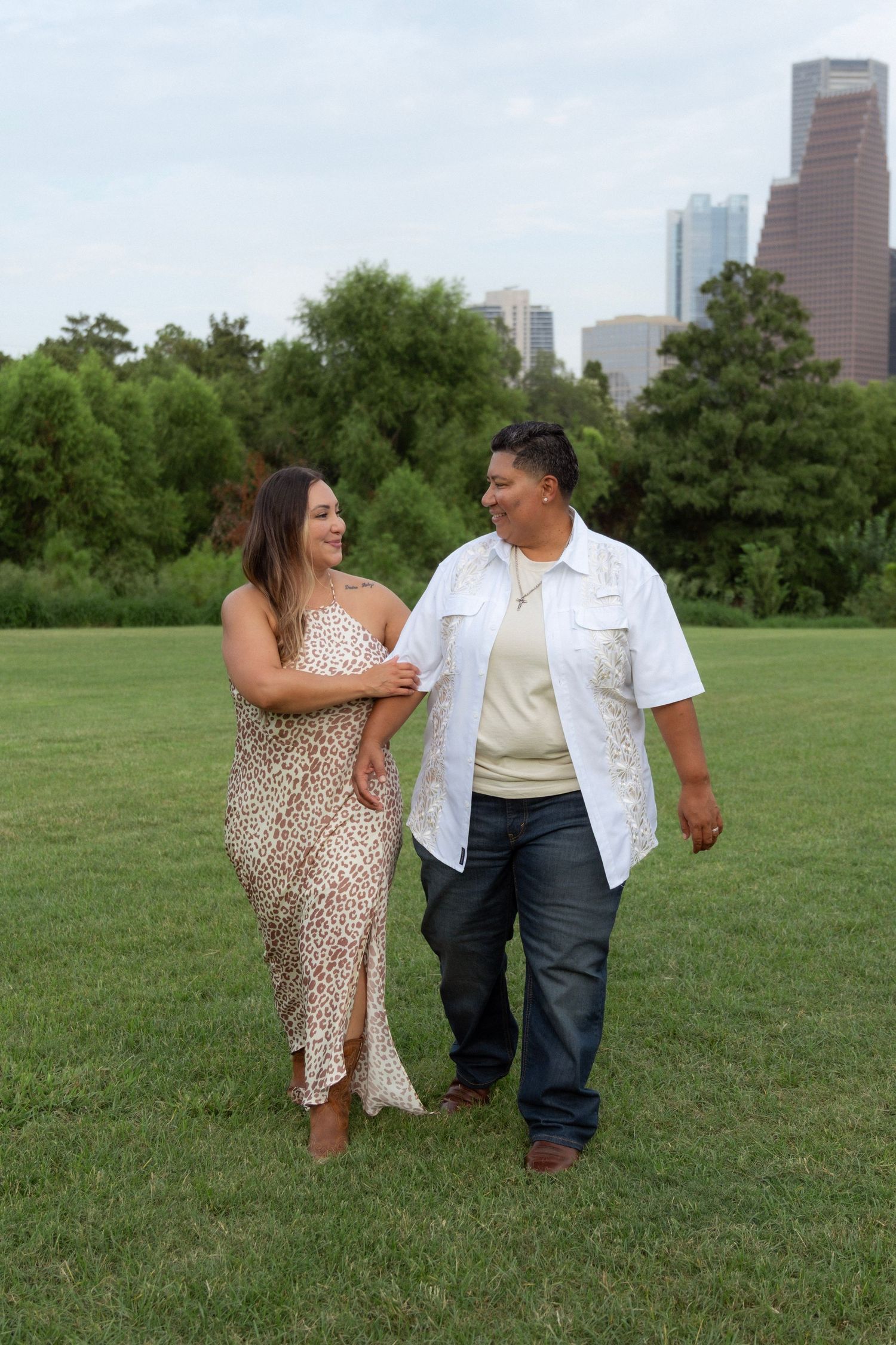 Couple walking hand in hand on green grass with city skyline and trees in background on a cloudy day.