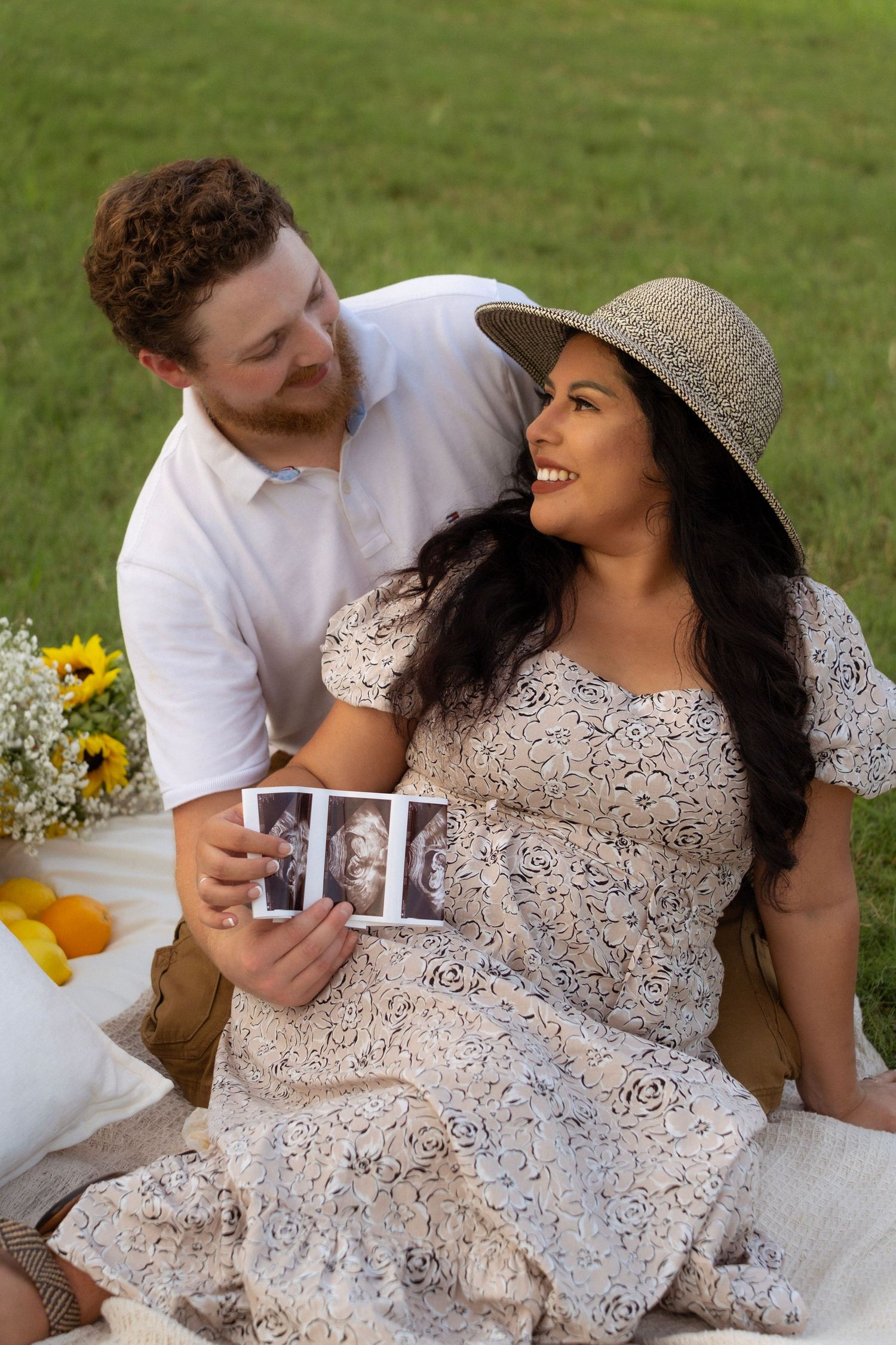 Happy couple sitting on grass sharing ultrasound photos during outdoor maternity picnic shoot.
