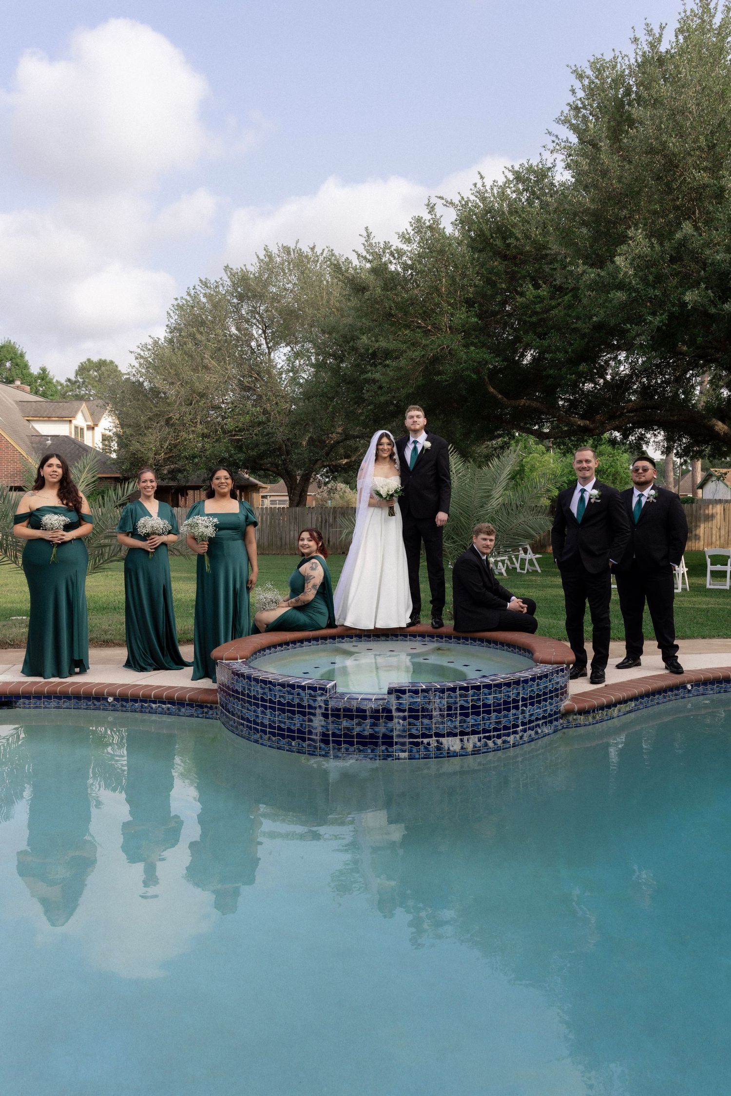 Wedding party poses on a fountain beside a pool with bride and groom in the center.