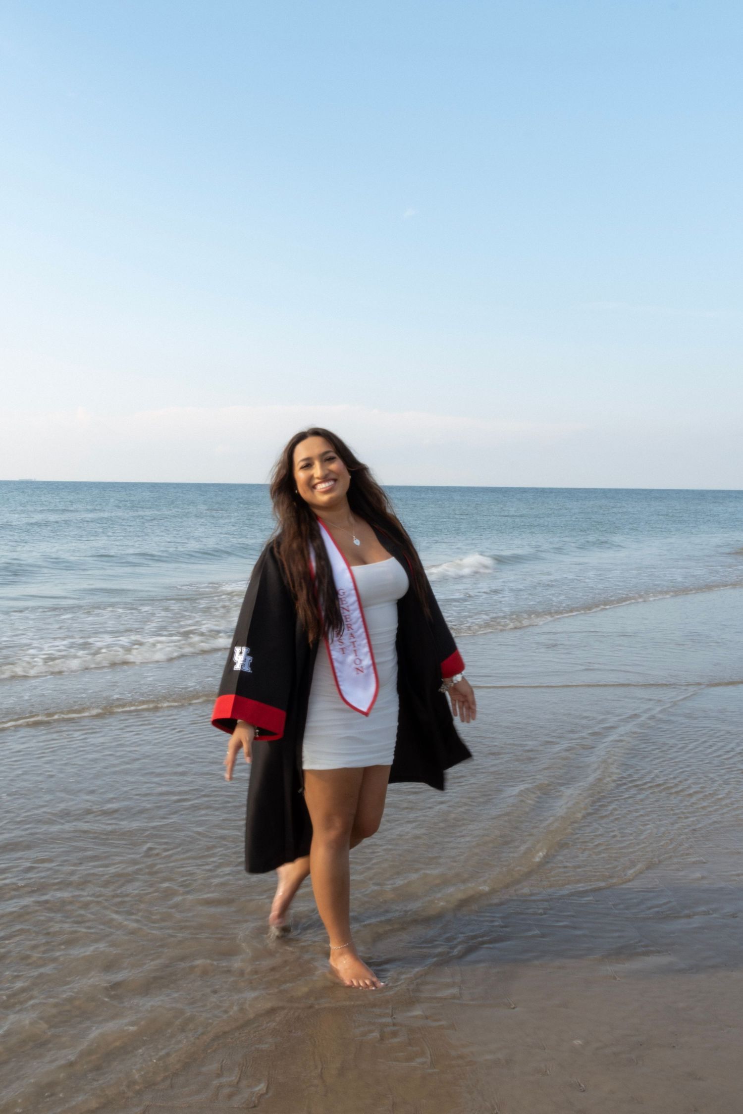 Smiling graduate in cap and gown walks barefoot along ocean shoreline on a clear sunny day.