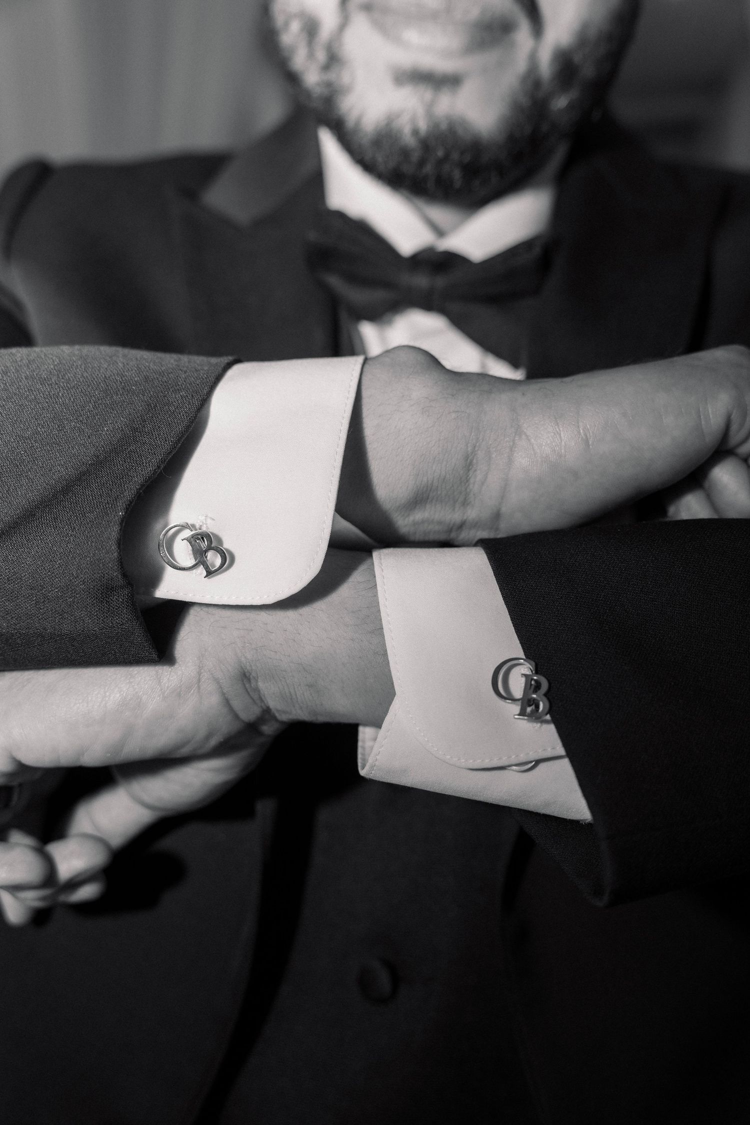 Black and white close-up of groom adjusting monogrammed cufflinks on tuxedo shirt with bow tie.