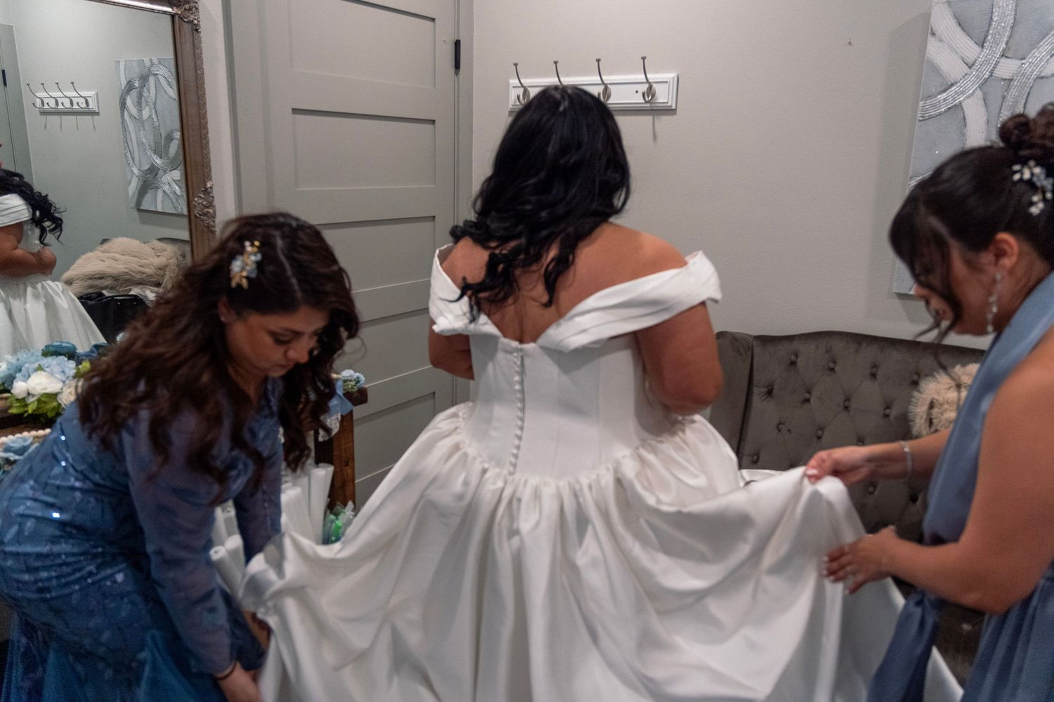 Two bridesmaids in blue dresses helping bride into elegant white off-shoulder wedding gown.