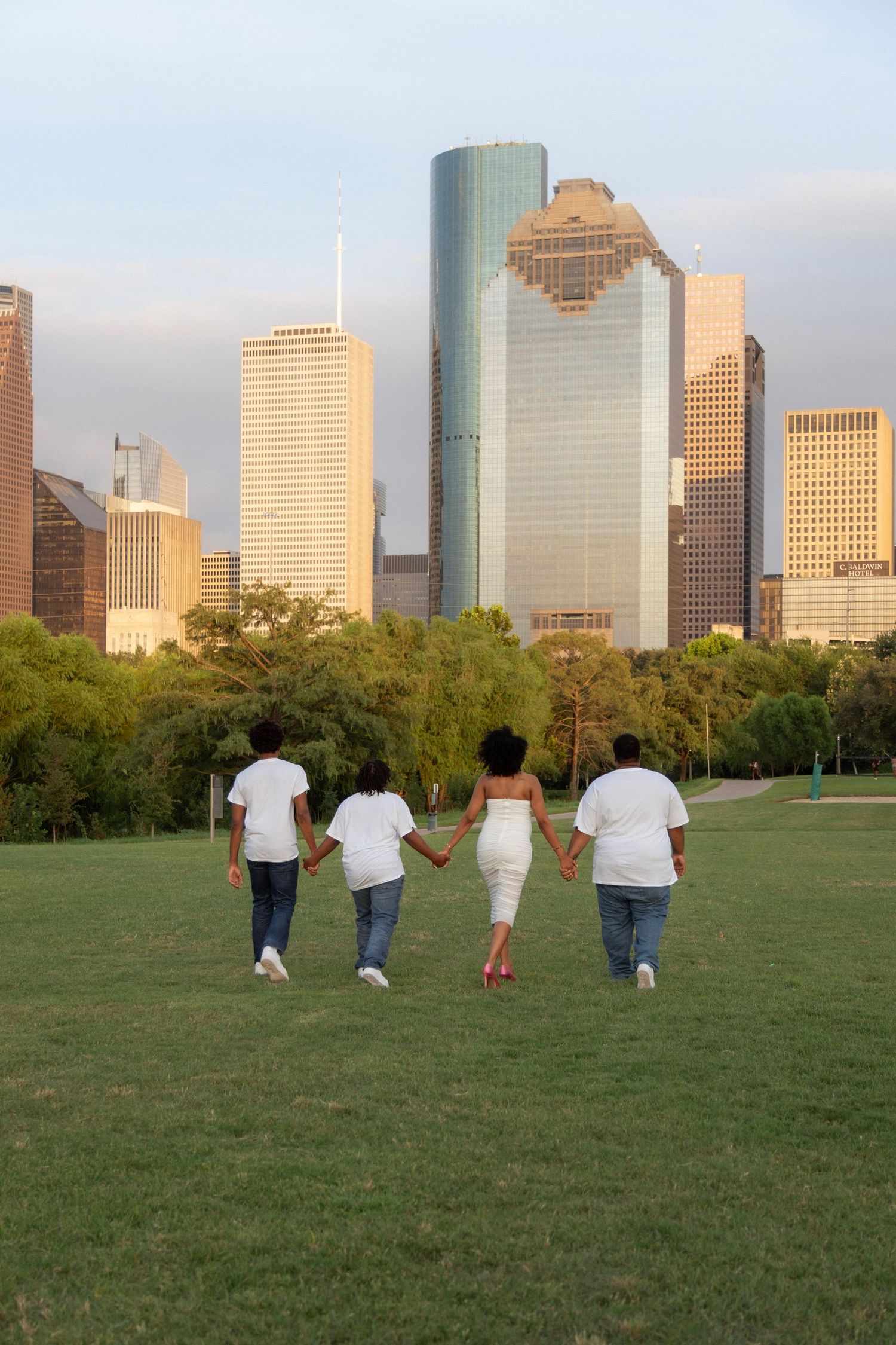 Family of four holding hands walking toward city skyline in a green park during golden hour.