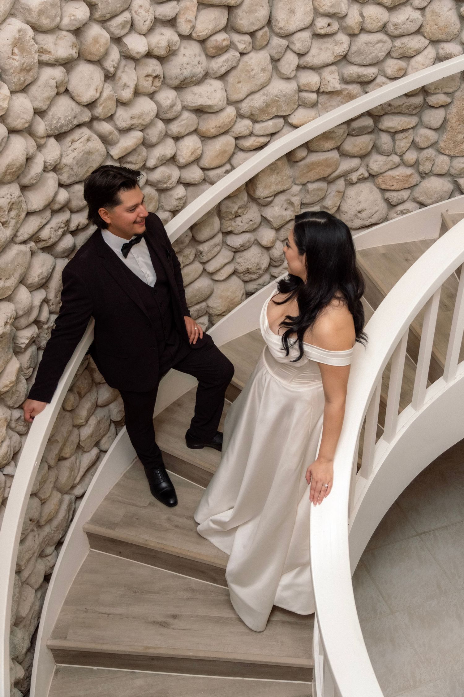 Elegant bride and groom share a glance on a curved white staircase with stone wall backdrop.