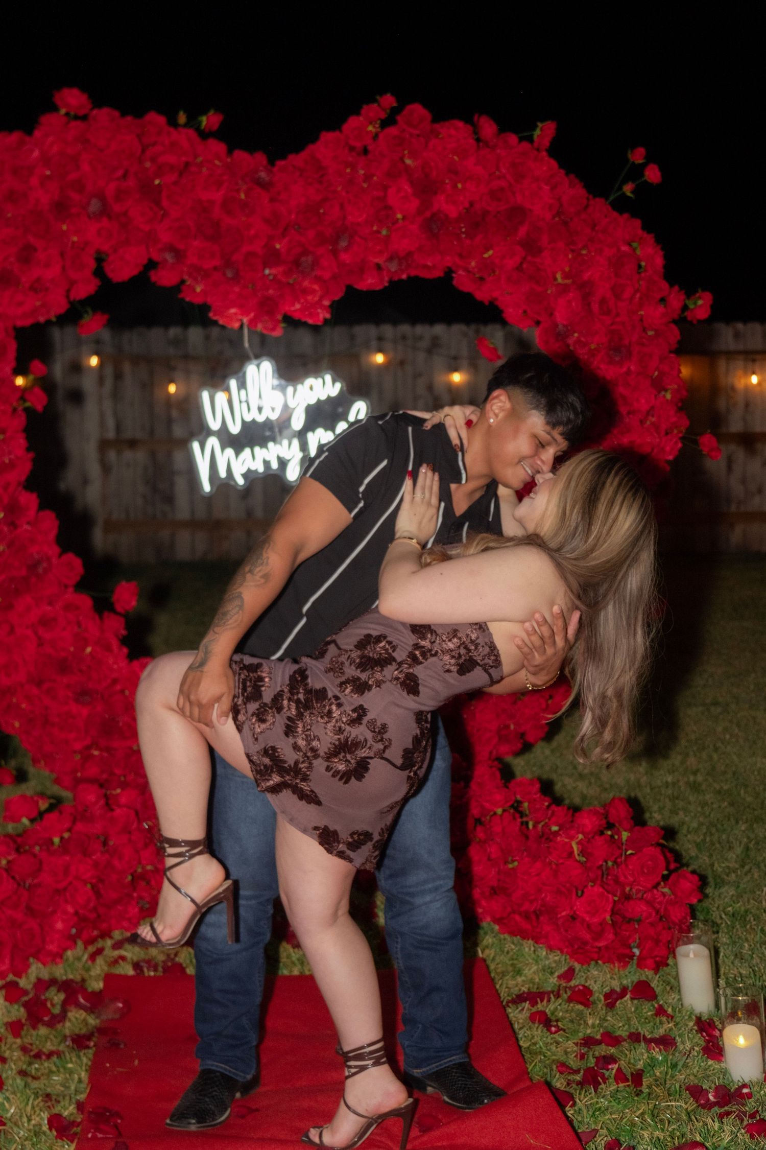 Couple sharing passionate kiss in front of heart-shaped red rose arch with Will You Marry Me sign.